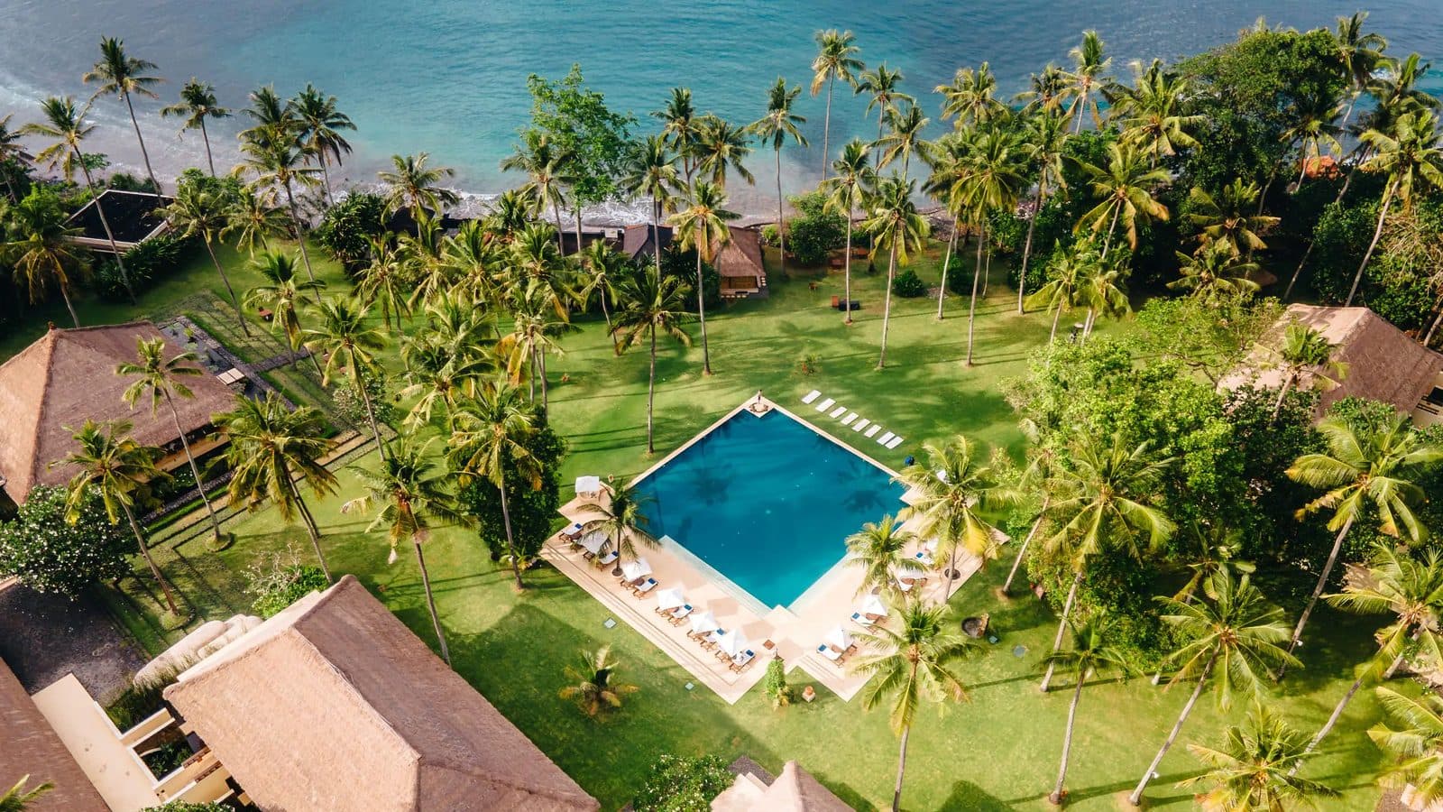Alila Manggis from above: a square pool on a lawn among coconut palms, roofed pavilions and a strip of beach by the sea