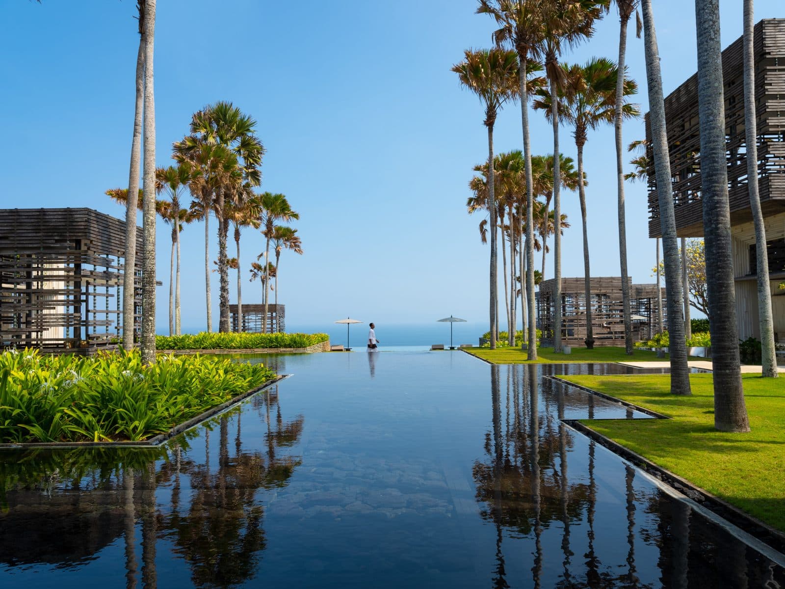 Alila Villas Uluwatu: длинный зеркальный бассейн между газонами и высокими пальмами уходит к горизонту океана, по краям решётчатые деревянные павильоны