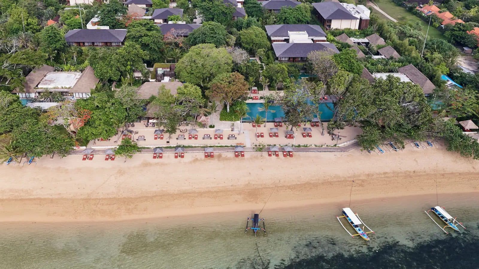 Вид сверху на Andaz Bali и пляж Санура: крыши среди деревьев, бассейн у берега, ряды лежаков и лодки-джукунги на мелководье