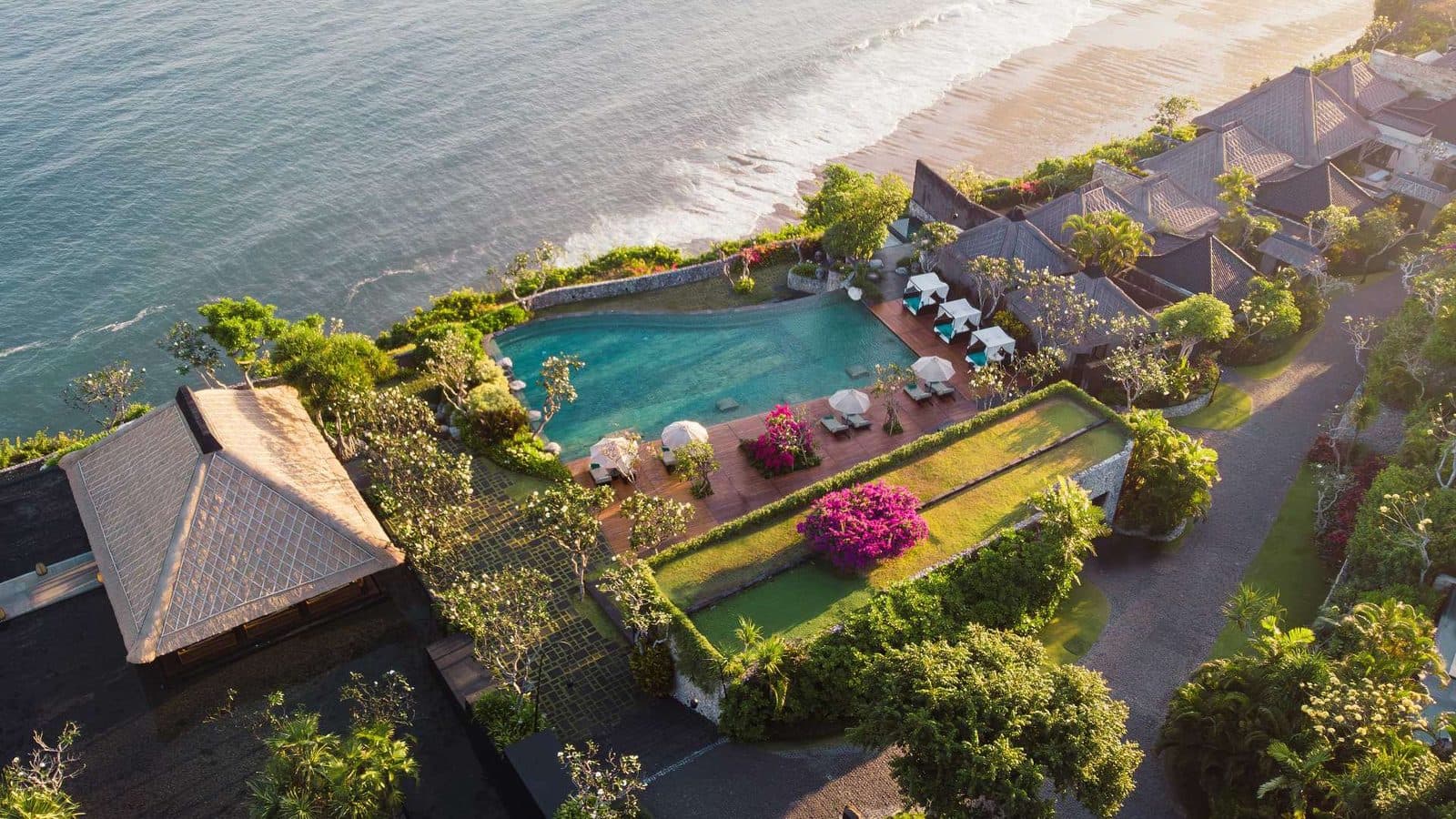 Вид сверху на Bvlgari Resort Bali на закате: главный бассейн и лужайки на краю утёса, внизу пляж и океан