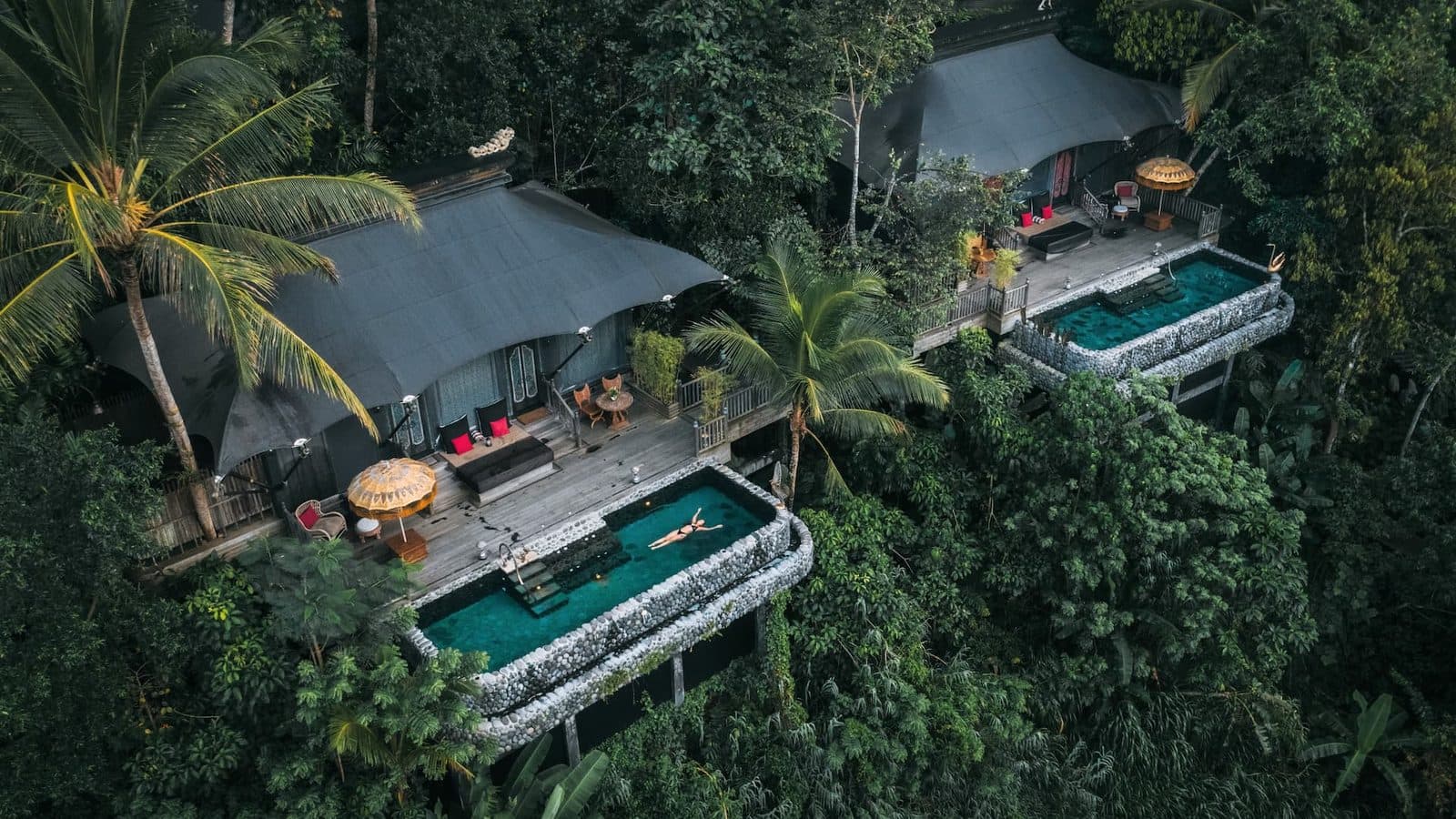 Aerial view of two Capella Ubud tents in the forest: wooden terraces, umbrellas and stone-edged pools above the slope