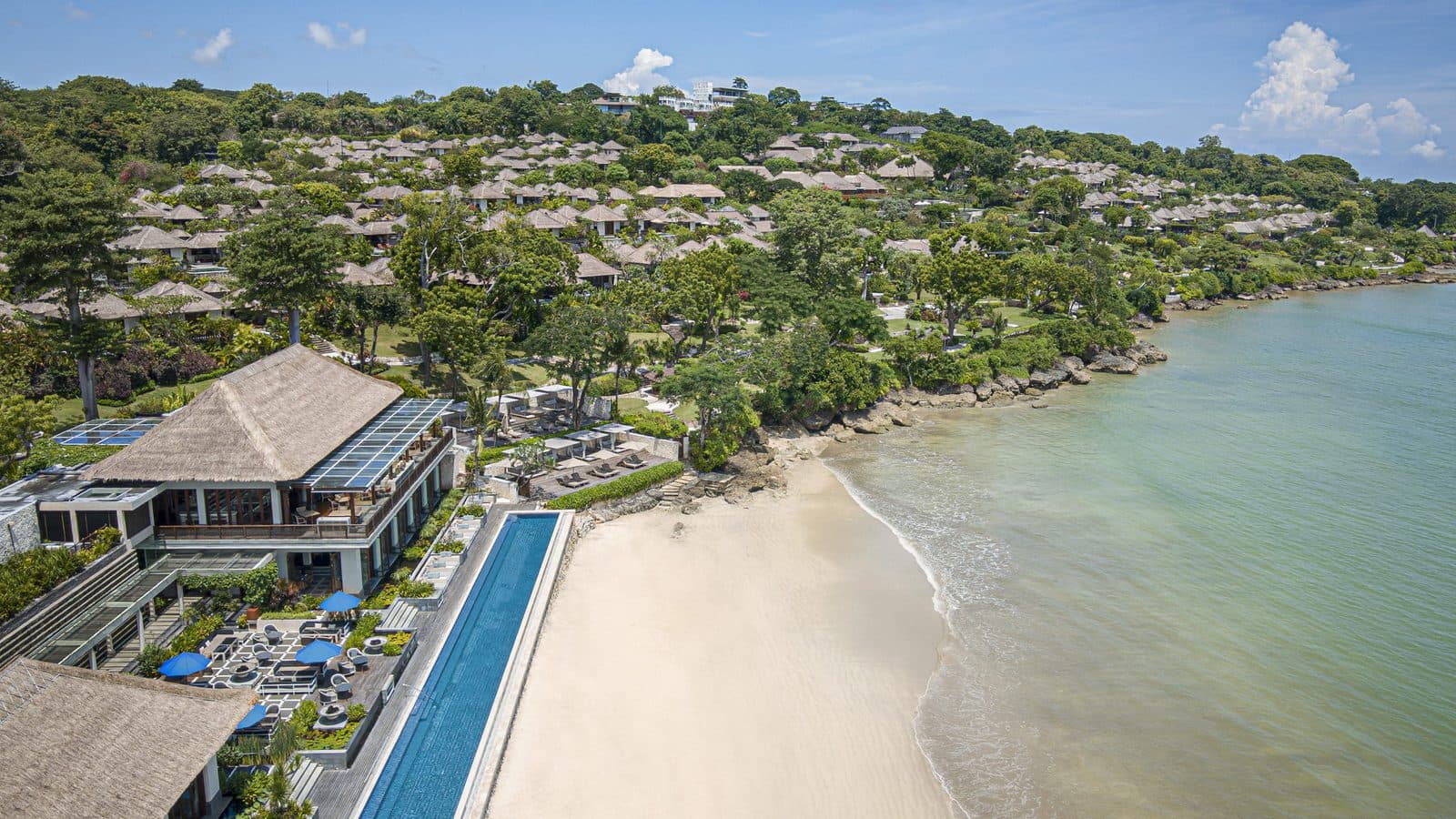 Вид с воздуха на Four Seasons Jimbaran Bay: виллы под соломенными крышами на склоне, длинный бассейн Sundara вдоль пляжа и залив
