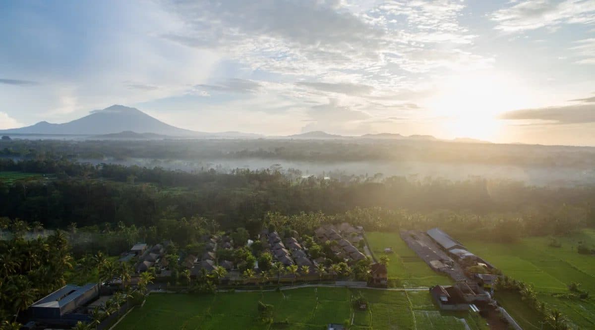 Вид сверху на HOSHINOYA Bali на рассвете: крыши вилл среди пальм и рисовых полей, туман над долиной и вулкан на горизонте