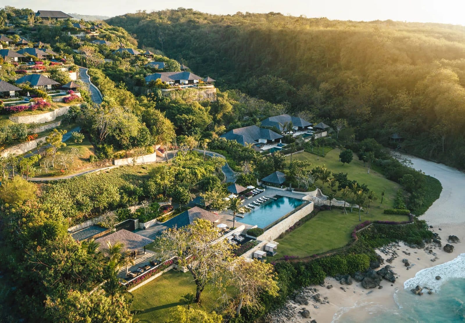 Вид сверху на Raffles Bali: виллы под тёмными крышами на зелёном склоне, бассейн у газона и пляж в бухте под лесистым холмом