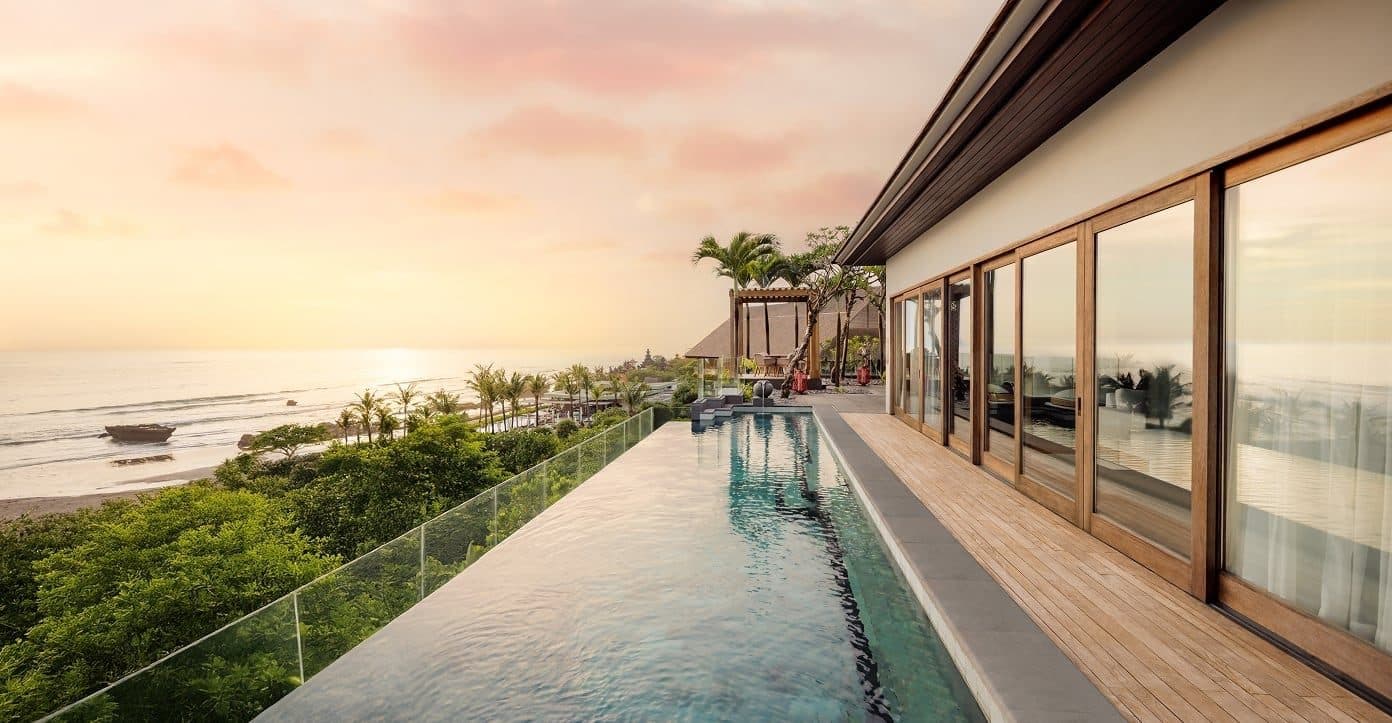 Бассейн-инфинити пентхауса Regent Bali Canggu на закате: длинная чаша вдоль стеклянной стены, внизу пальмы и пляж с лодкой