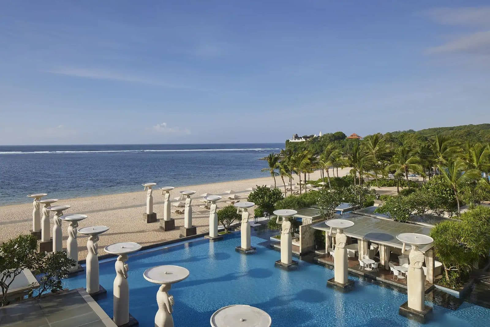 Бассейн-инфинити Oasis в The Mulia: белые статуи с чашами вдоль воды, за ними пляж Гегер, океан и мыс с пальмами