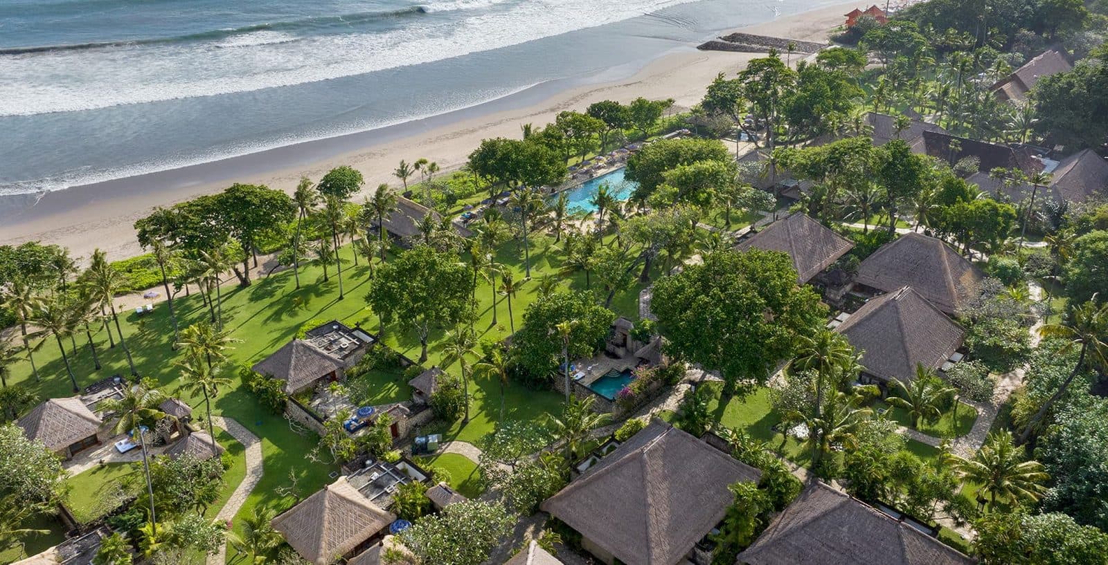 Вид сверху на The Oberoi, Bali: соломенные крыши вилл среди пальм и газонов, бассейн у кромки сада и пляж Семиньяка с прибоем