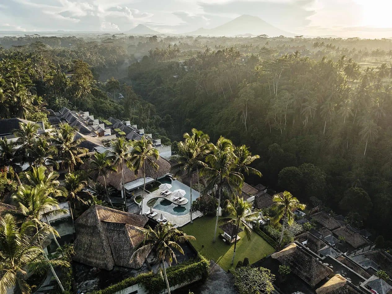 Вид сверху на Viceroy Bali на рассвете: виллы под соломенными крышами, круглый бассейн с зонтами и лужайка над долиной в джунглях, вдали вулкан