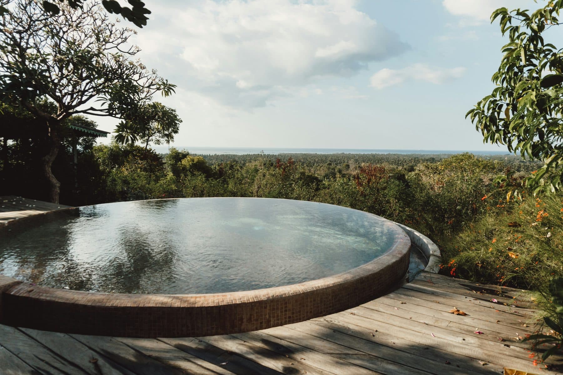 A circular infinity pool on a villa's wooden terrace above Bali's green forest, with a strip of sea in the distance