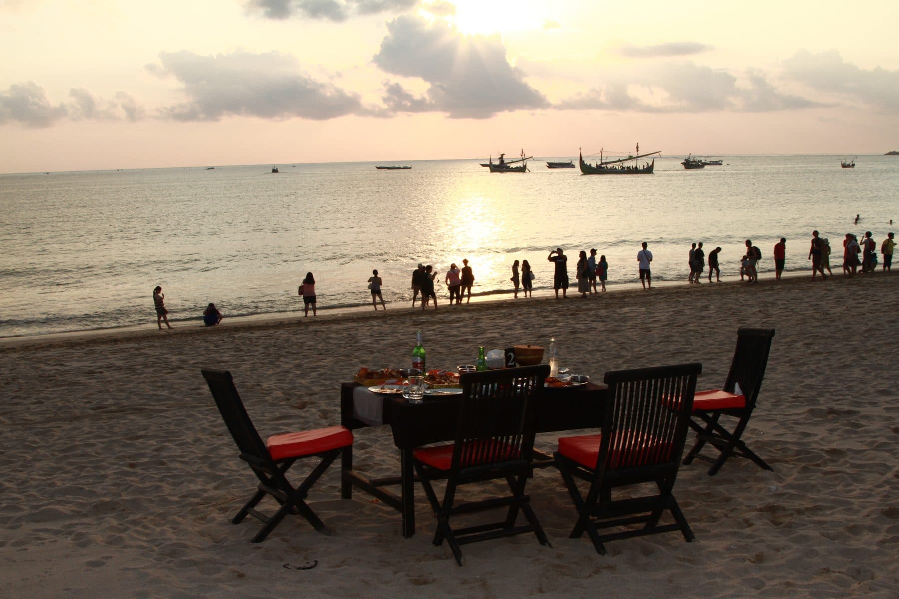 A table set for dinner and chairs with red cushions on Jimbaran Beach at sunset; people walk by the water and fishing boats lie in the bay