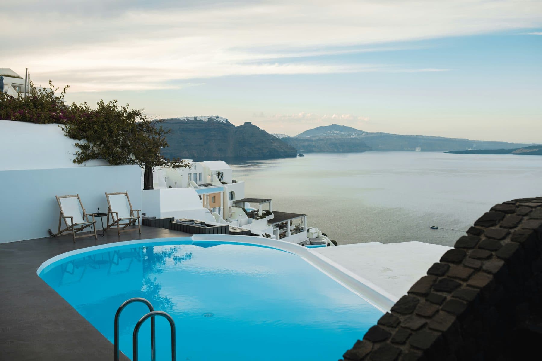 A hotel pool on the edge of Santorini’s caldera: two loungers on the terrace, white houses down the slope and the evening sea with islands
