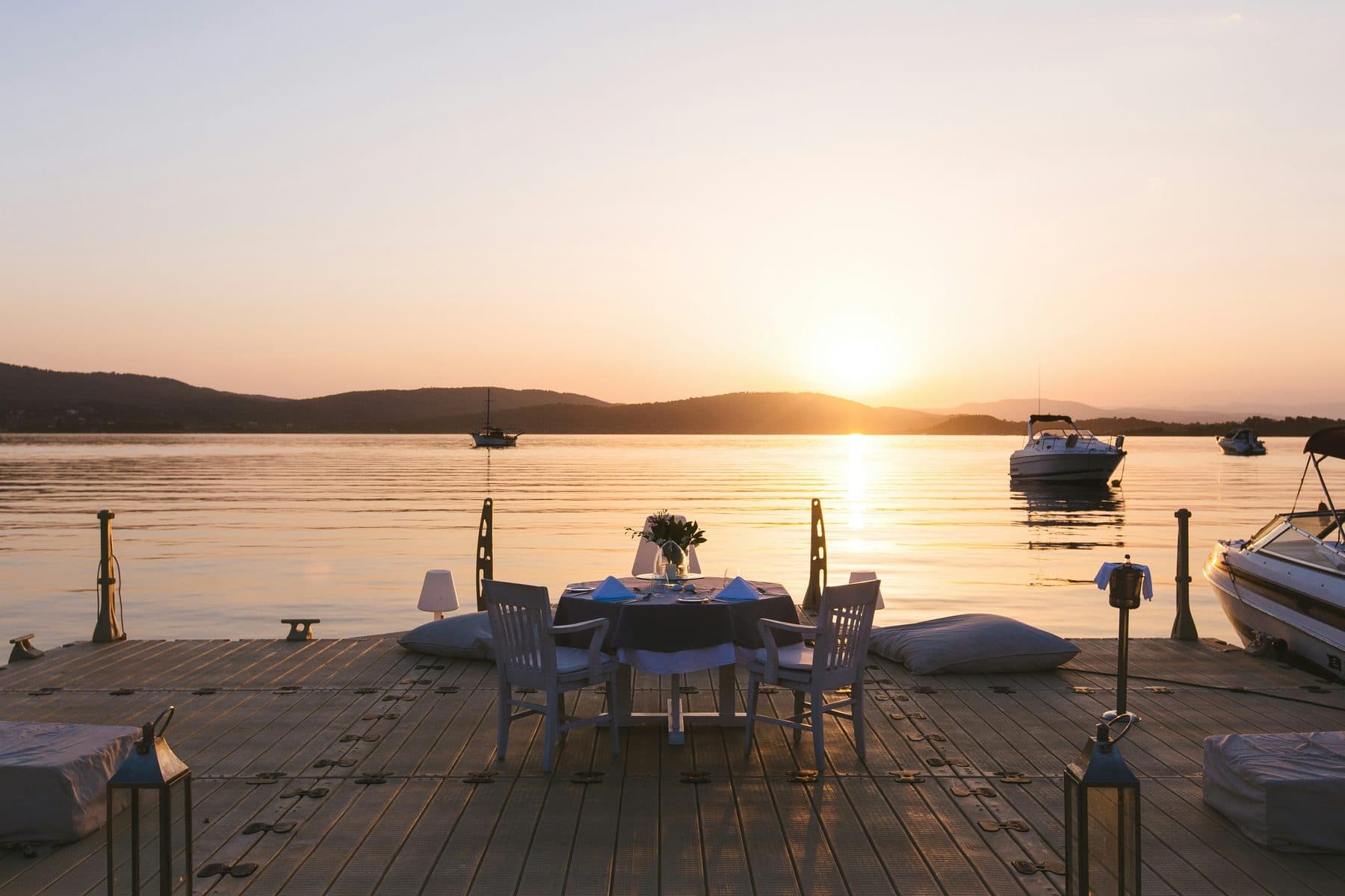 A table for two on a wooden jetty beside the sea in Greece at sunset, with lanterns and cushions nearby, boats and a sailing yacht in the bay