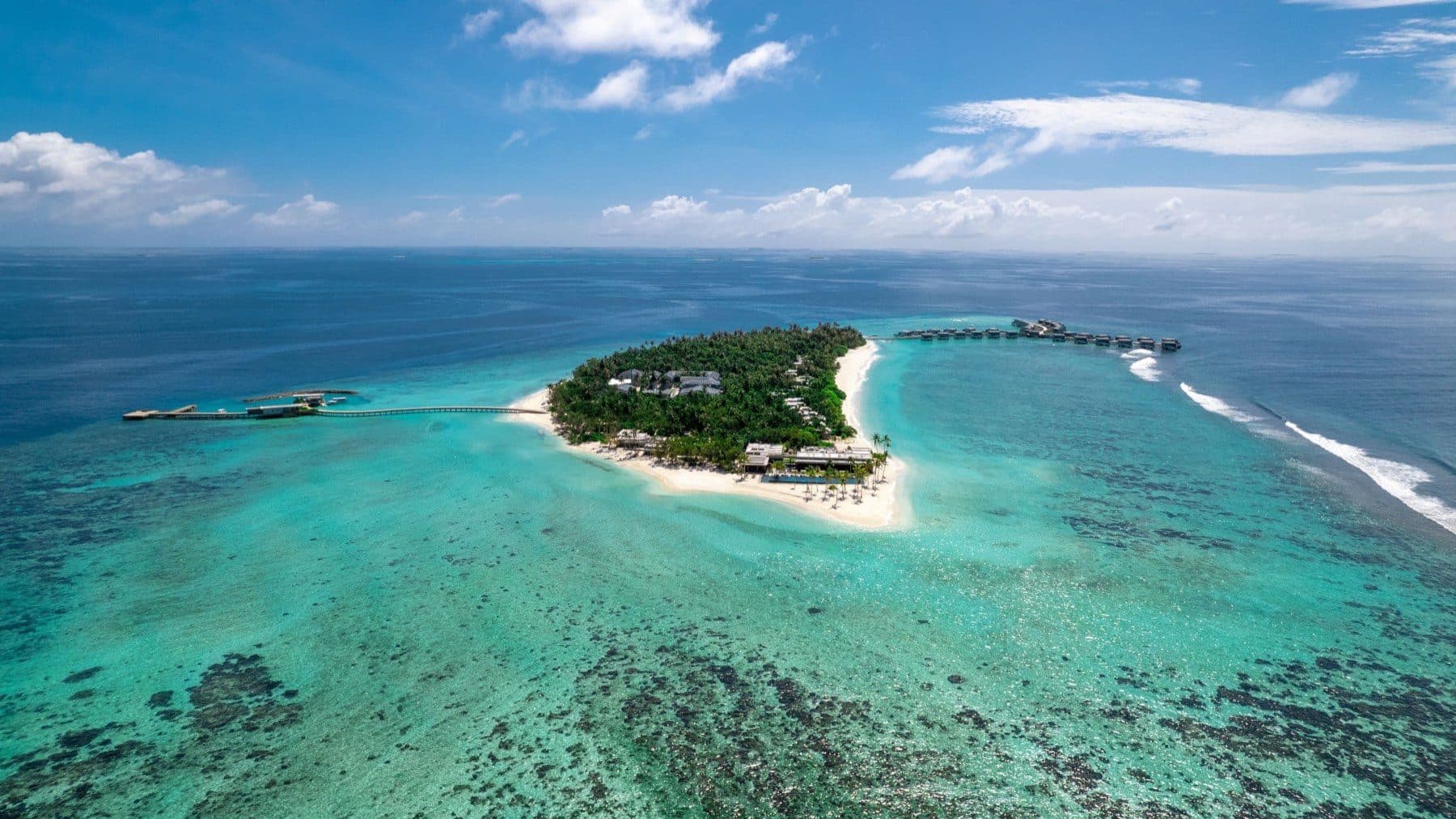 A small palm-covered island with a beach amid the reef, viewed from above: a long jetty on the left and a row of overwater villas on the right