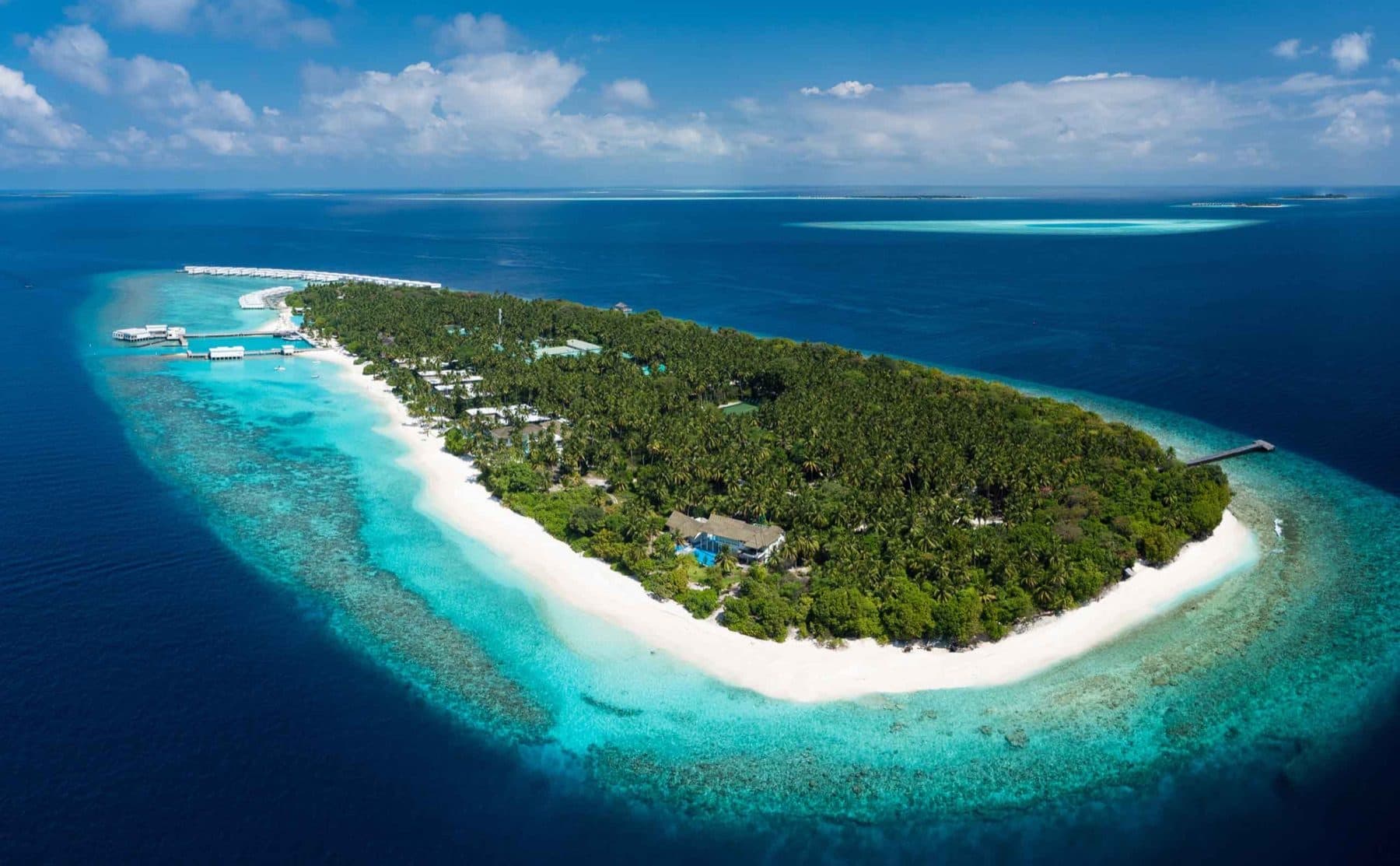 Aerial view of an island with dense palms and a white beach inside a ring of reef; a row of overwater villas stands off the shore on the left