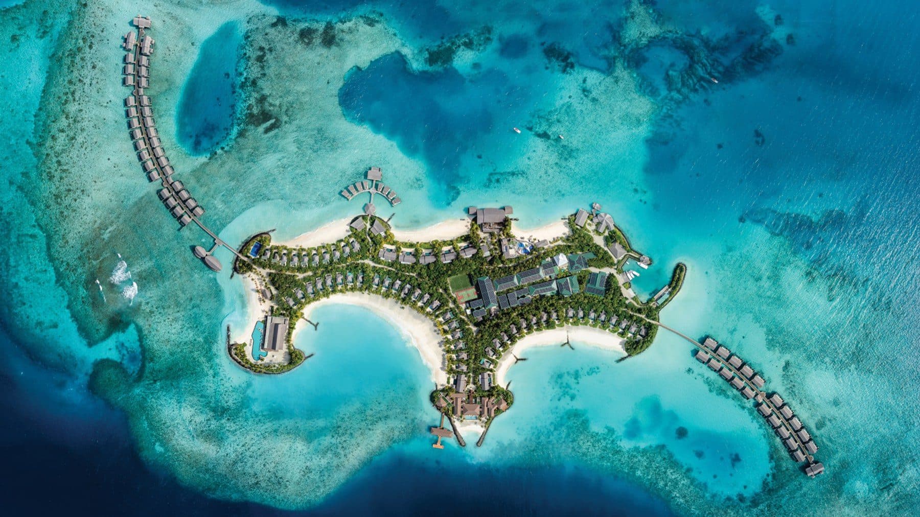Direct overhead view of an island with beach villas among greenery and two rows of overwater villas on walkways above the reef