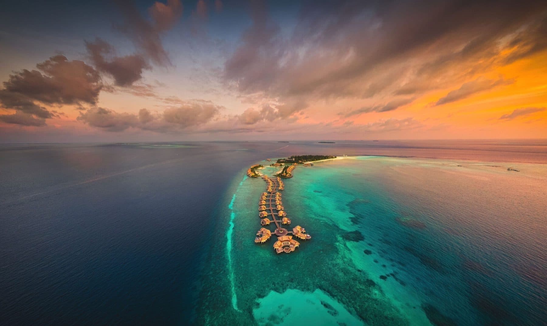 An island and a long chain of overwater villas on walkways above a turquoise lagoon at sunset, viewed from above