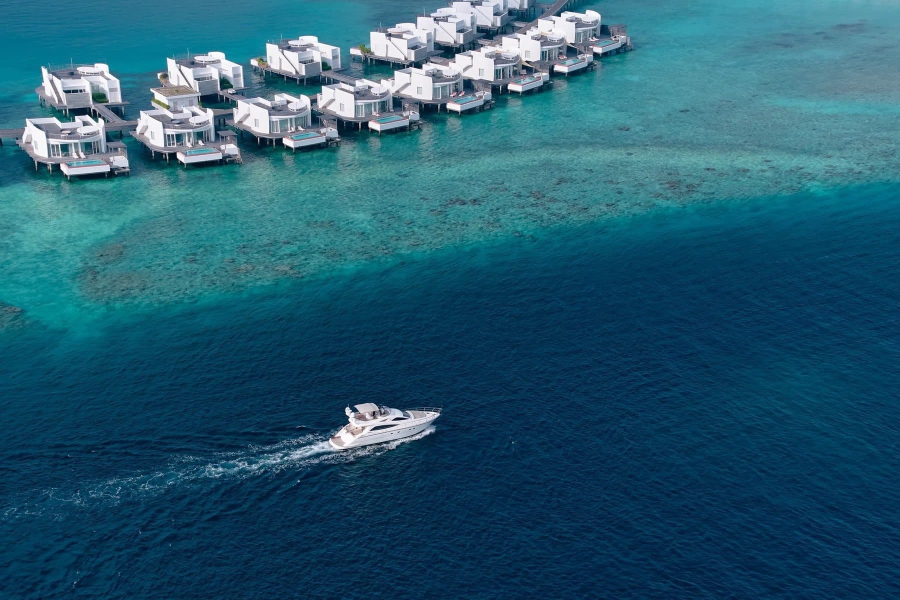 White overwater pool villas on stilts above the reef and a yacht out at sea, viewed from the air