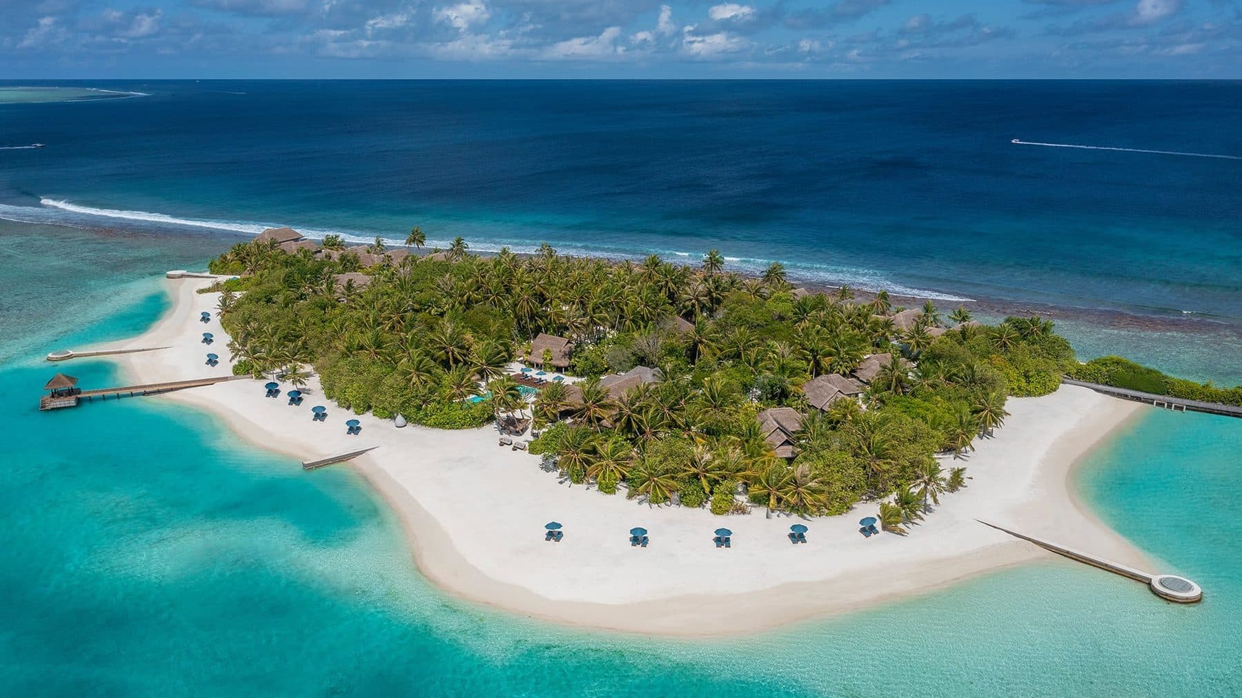 A palm-covered island with a wide white beach, umbrellas along the shore, a lagoon and reef, viewed from the air