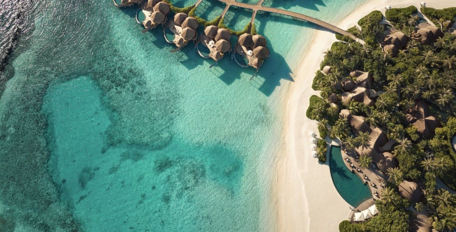 Aerial view of overwater houses on stilts, a white beach, a pool beside the restaurant and the island's palms