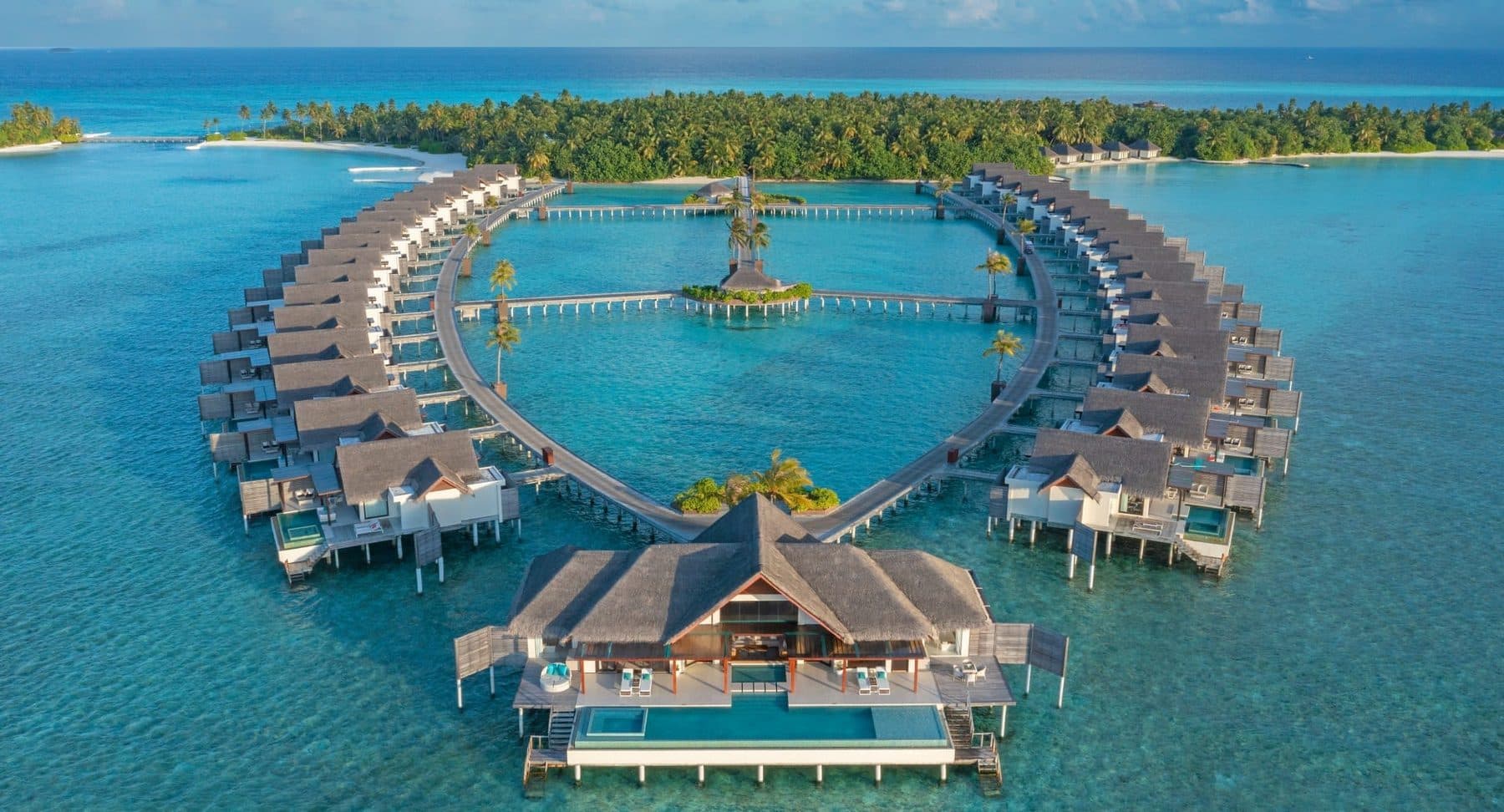 A ring of stilt villas around a lagoon, with a palm-covered island behind, viewed from the air