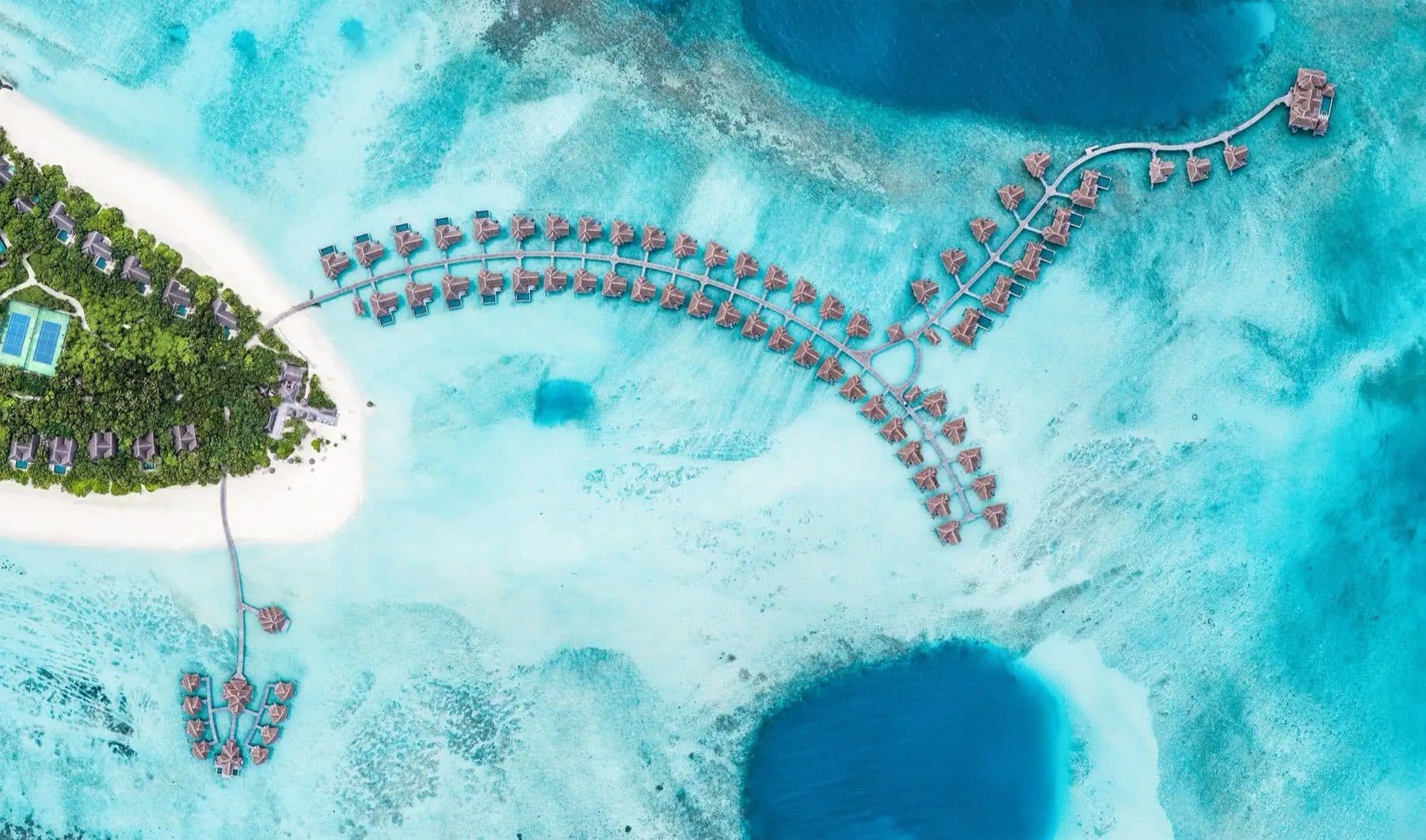 Direct overhead view of the island's edge with villas and tennis courts, and long walkways with overwater villas above the lagoon between two deep blue depressions