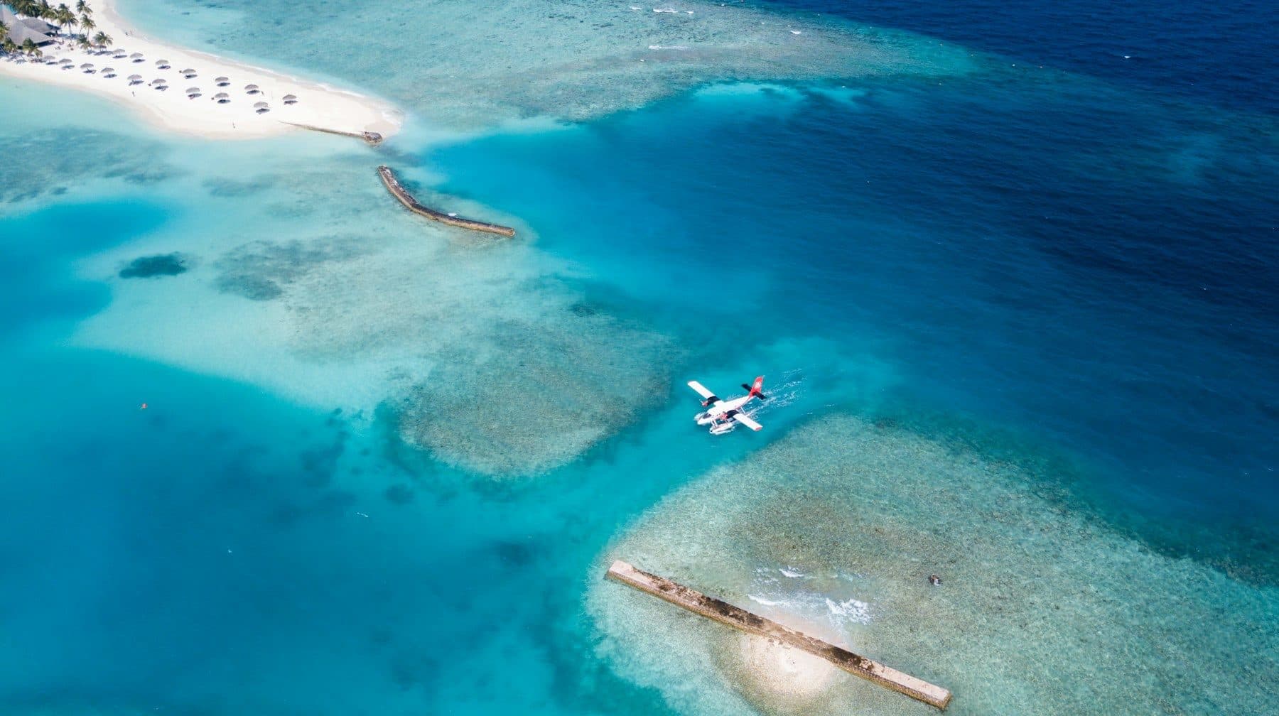 A seaplane on the water by the reef near Veligandhu Island, viewed from above; a beach with umbrellas on the left.