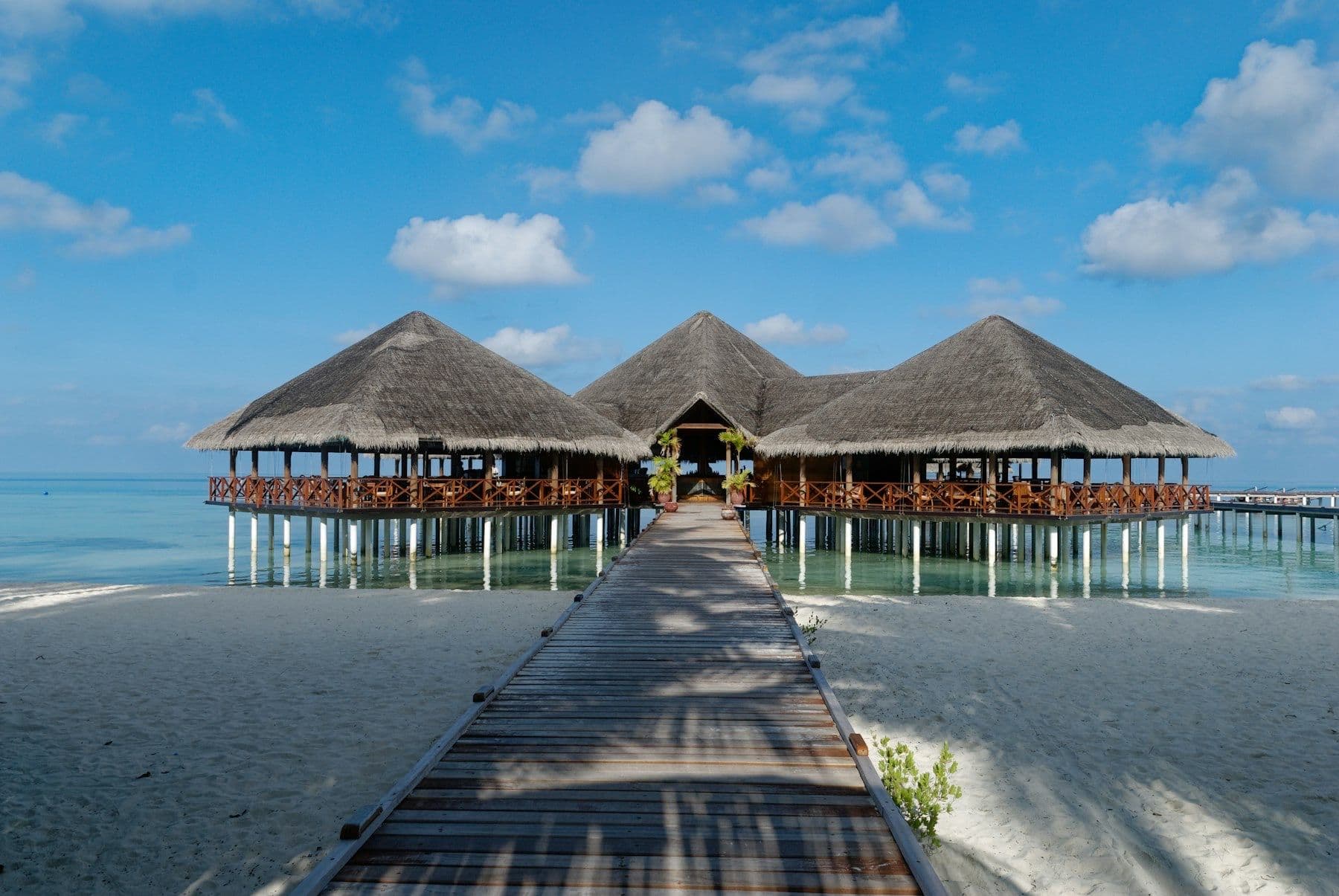 Wooden walkways across white sand lead to a thatched pavilion on stilts above the lagoon.