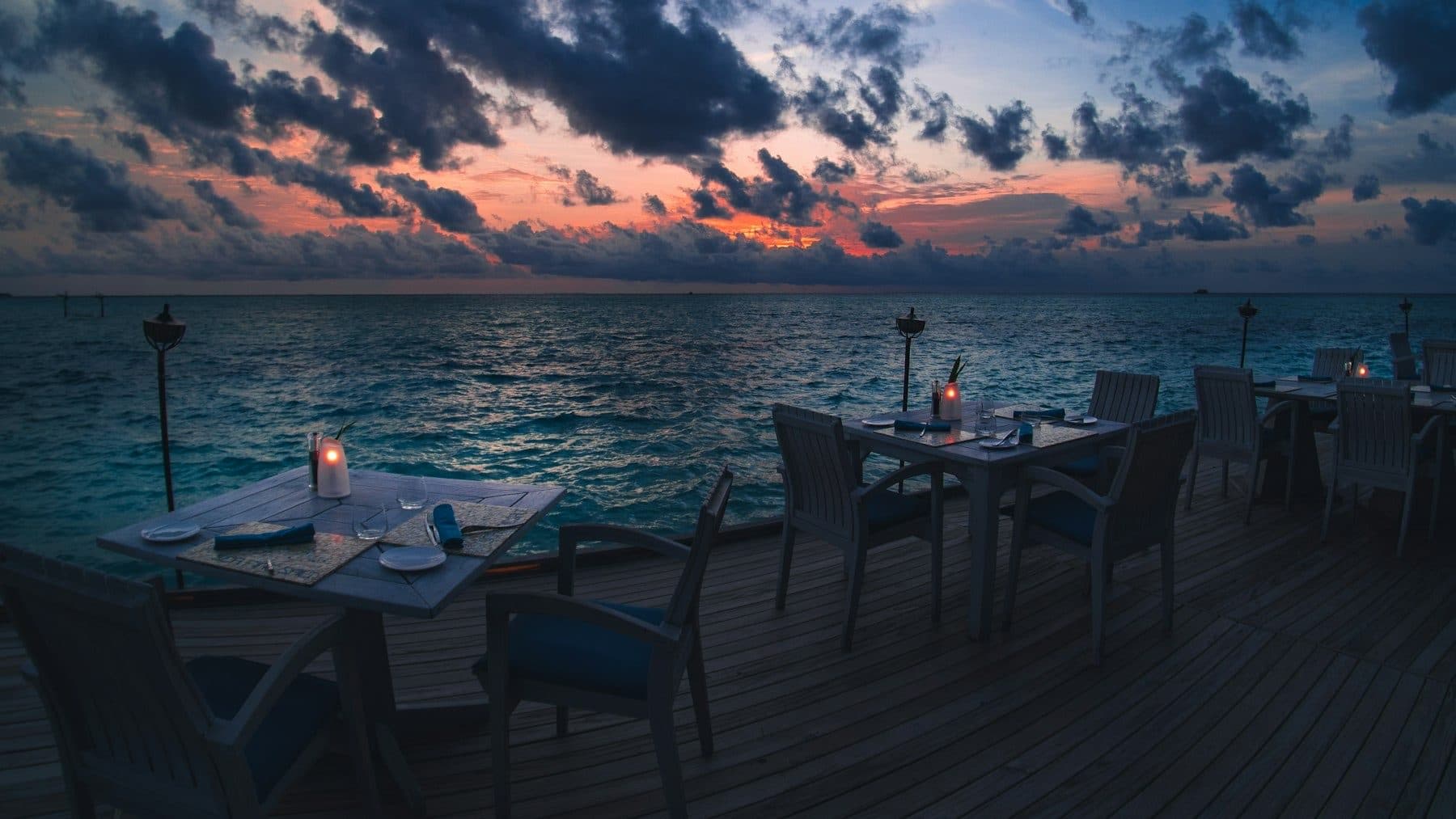 An overwater restaurant terrace at sunset: candlelit tables and torches along the edge of the wooden deck.