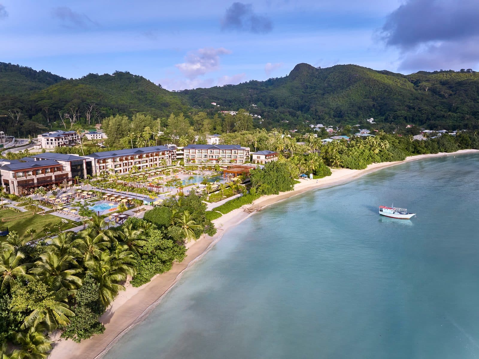 Вид сверху на Canopy by Hilton Seychelles: корпуса и бассейны среди пальм вдоль пляжа бухты Анс-а-ла-Муш, лодка на бирюзовой воде и зелёные горы Маэ