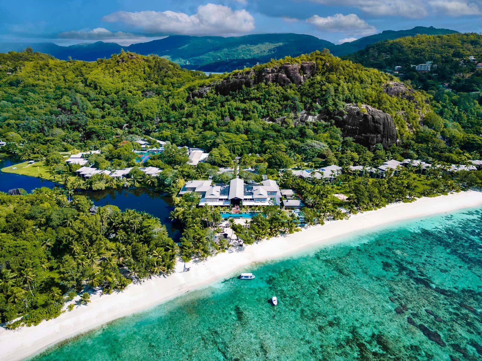 Вид сверху на Kempinski Seychelles: корпуса среди пальм между лагуной и пляжем бухты Бэ-Лазар, риф в бирюзовой воде, лесистые горы с гранитными скалами