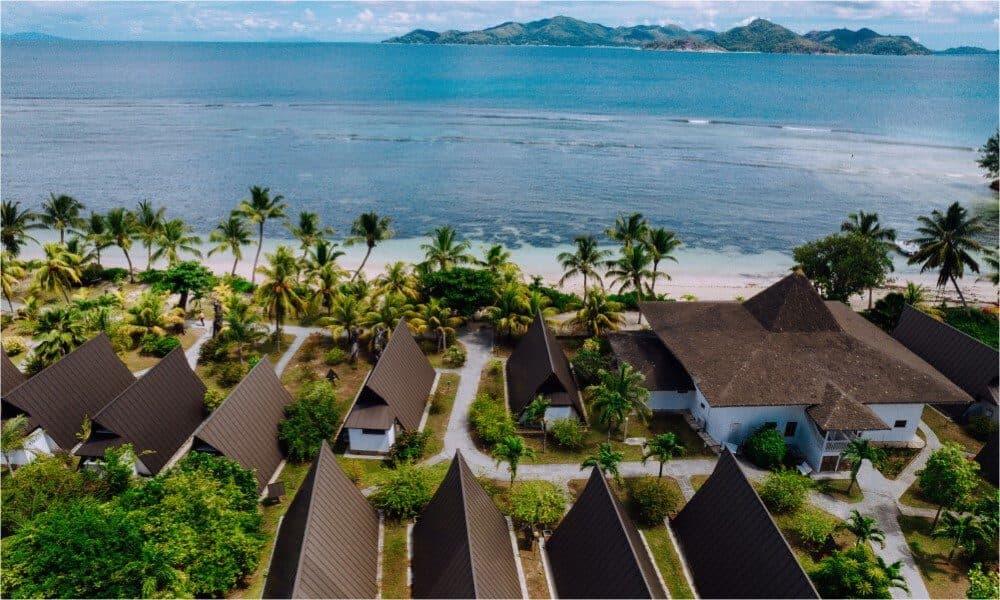 Вид сверху на La Digue Island Lodge: треугольные крыши шале среди пальм, белый песок пляжа Анс-Реюньон и Праслен за проливом