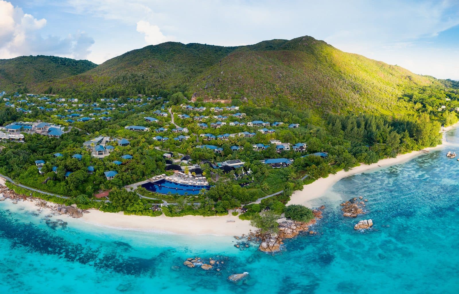 Вид сверху на Raffles Seychelles: виллы с синими крышами на зелёном склоне горы, большой бассейн у моря, белый пляж и бирюзовая вода с гранитными камнями