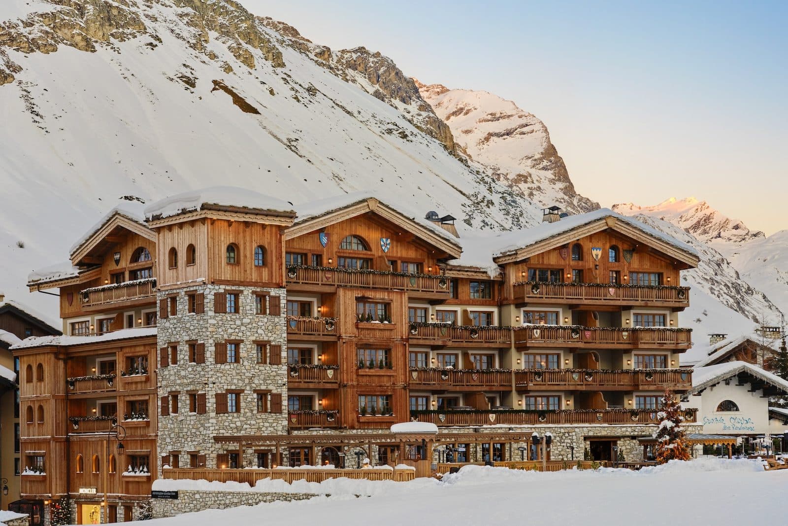 The snowy wood and stone façade of Airelles Val d’Isère, with a snowy slope and peaks in evening light behind