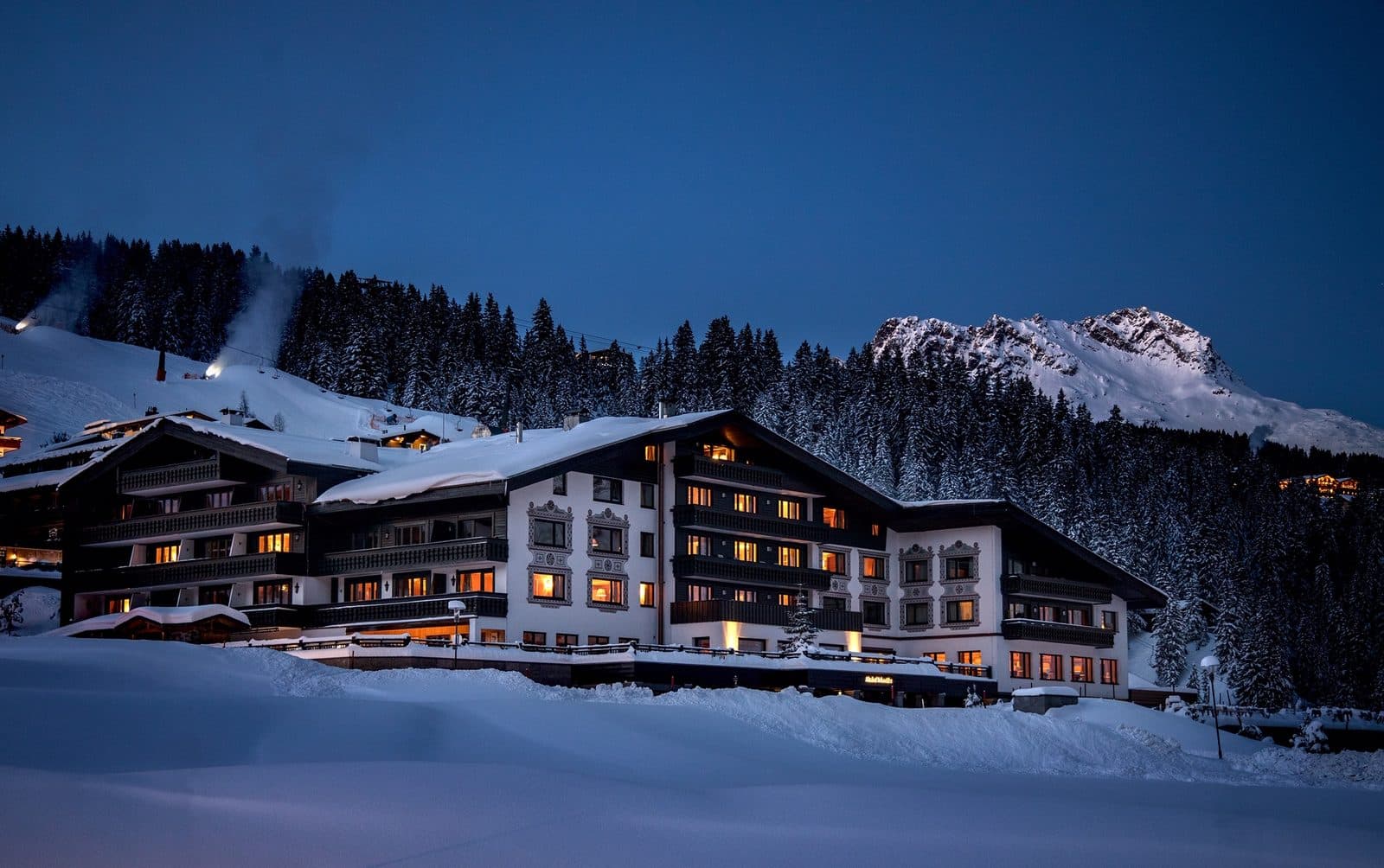 Illuminated Almhof Schneider buildings in the evening, surrounded by snow, conifer forest and mountains