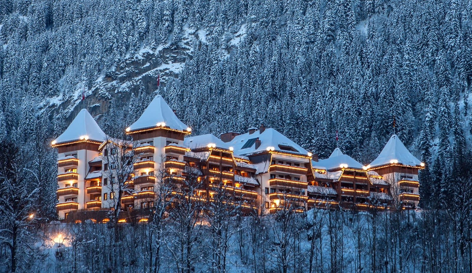 The Alpina Gstaad зимним вечером: здание с башенками под снежными крышами и светящимися балконами среди заснеженного елового леса