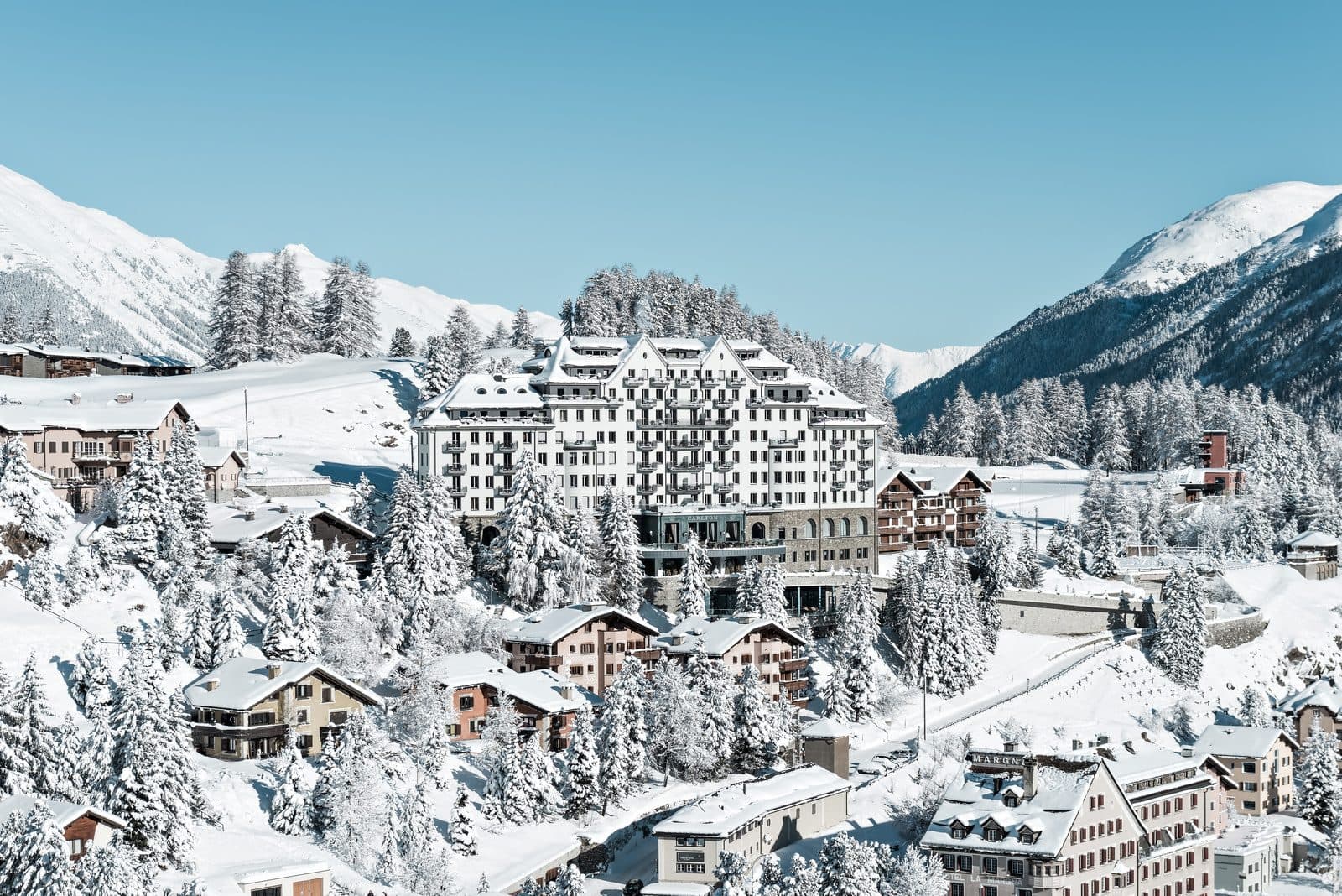 Carlton Hotel St. Moritz зимой: белое здание с башенками над заснеженными шале и елями, за ним горы Энгадина под синим небом
