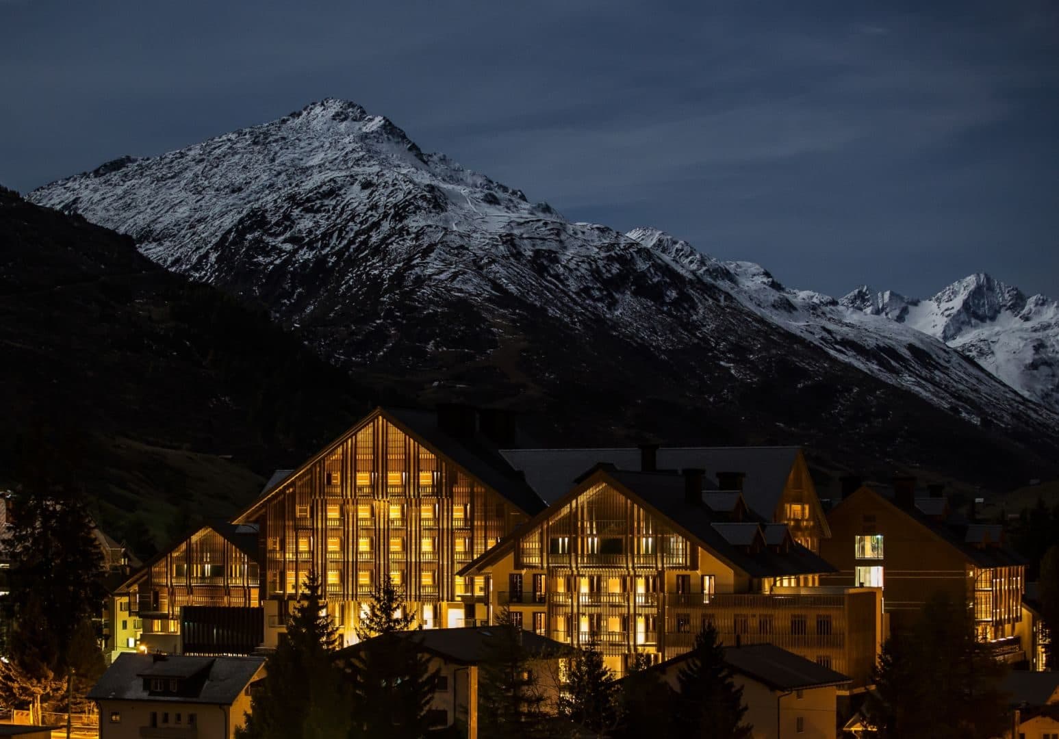 Освещённые корпуса The Chedi Andermatt вечером под заснеженной вершиной