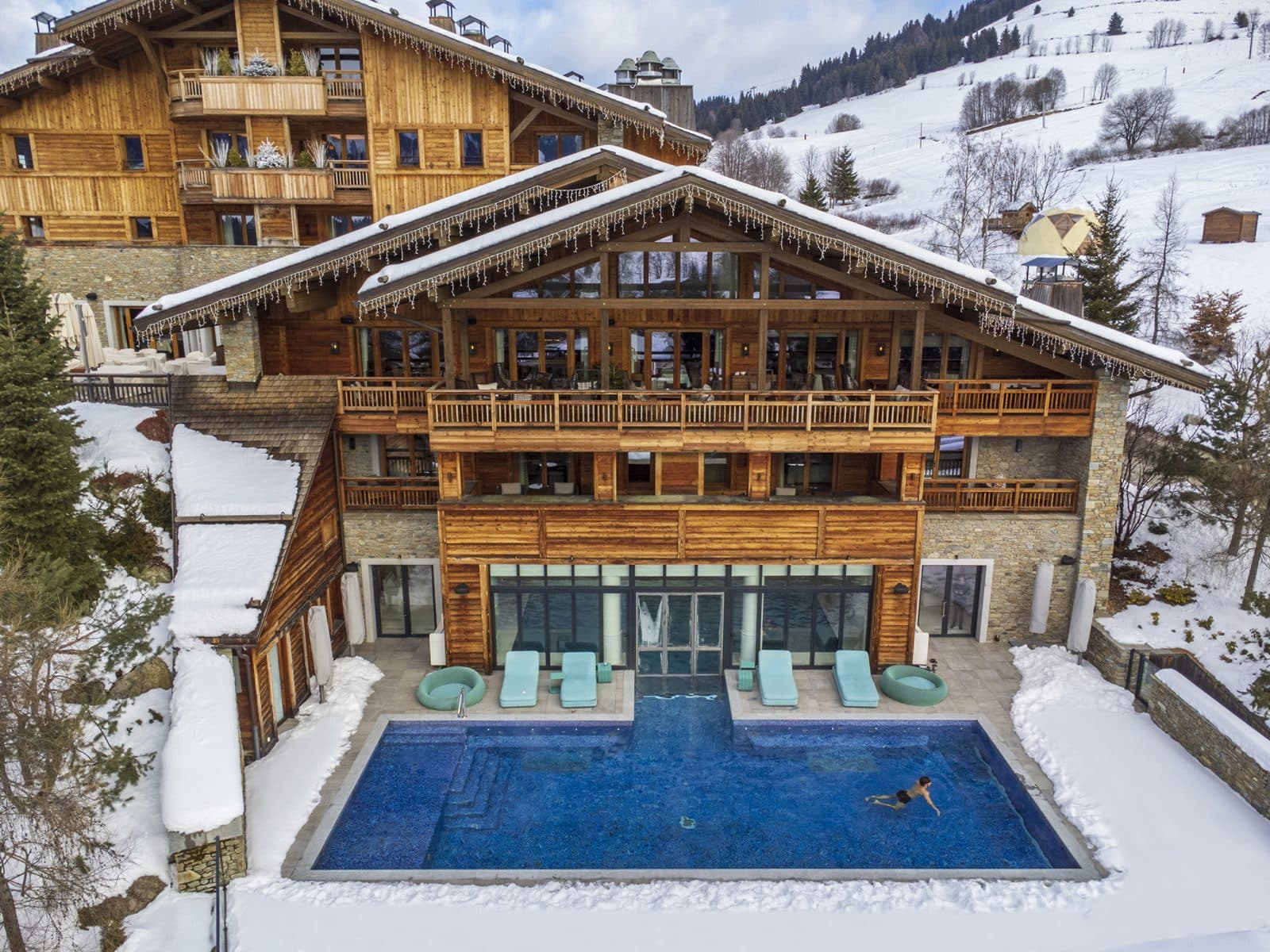 Вид сверху на деревянные шале Four Seasons Megève в снегу: открытый бассейн с бирюзовыми лежаками перед стеклянным фасадом спа