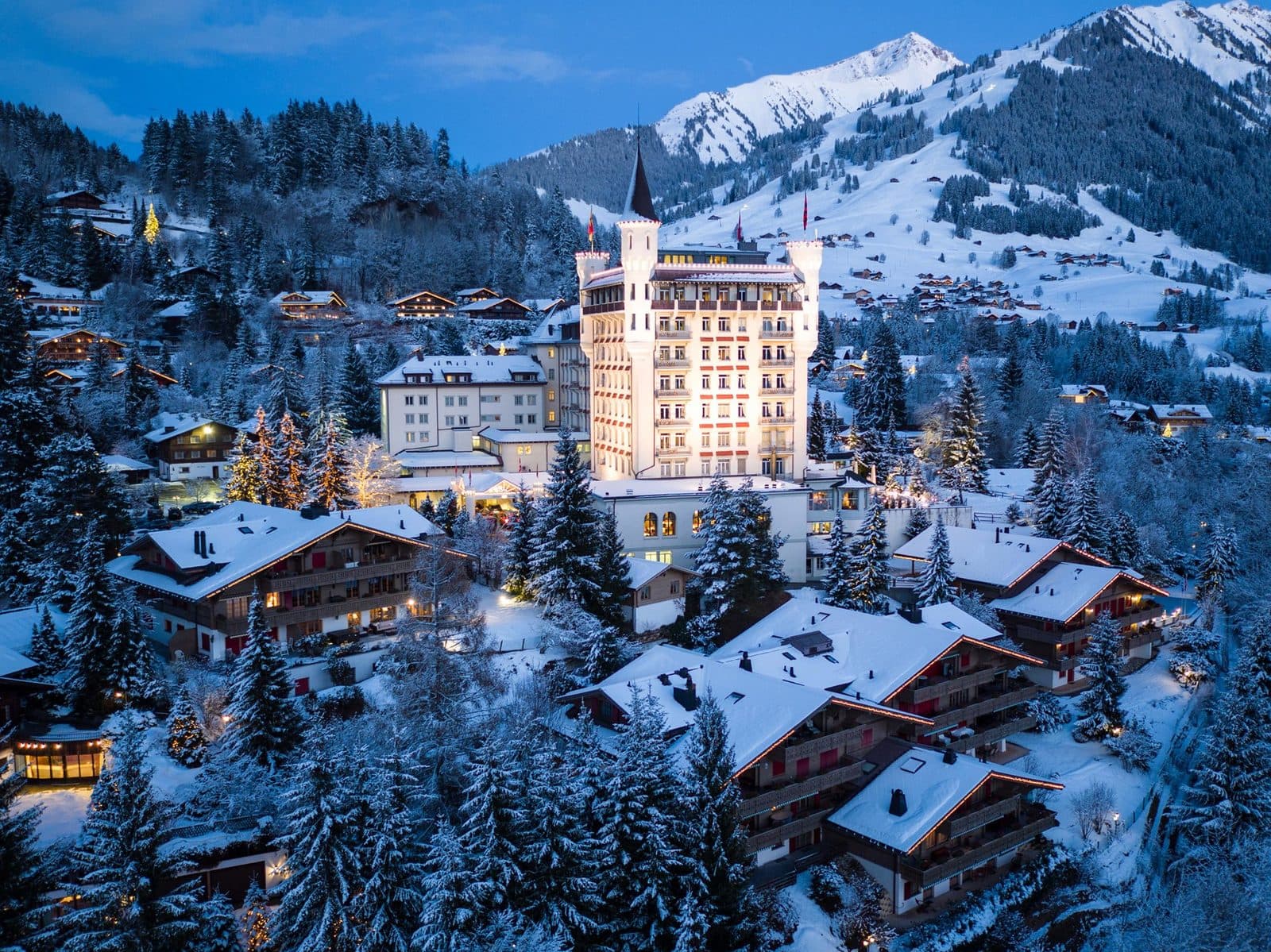 Gstaad Palace зимним вечером: белый замок с башнями над заснеженными шале Гштада, за ним склоны и вершины