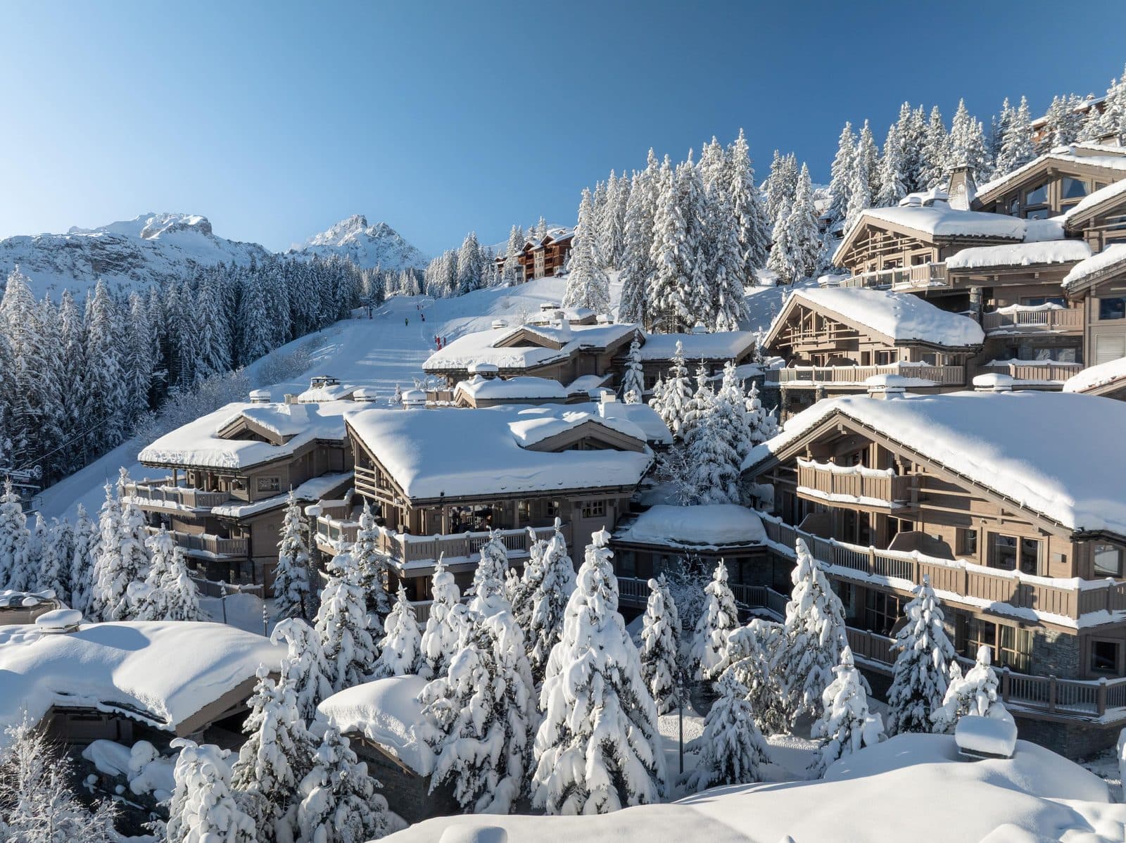Aerial view of Le K2 Palace: wooden and stone chalets beneath thick snow among snowy fir trees, with a piste and peaks behind