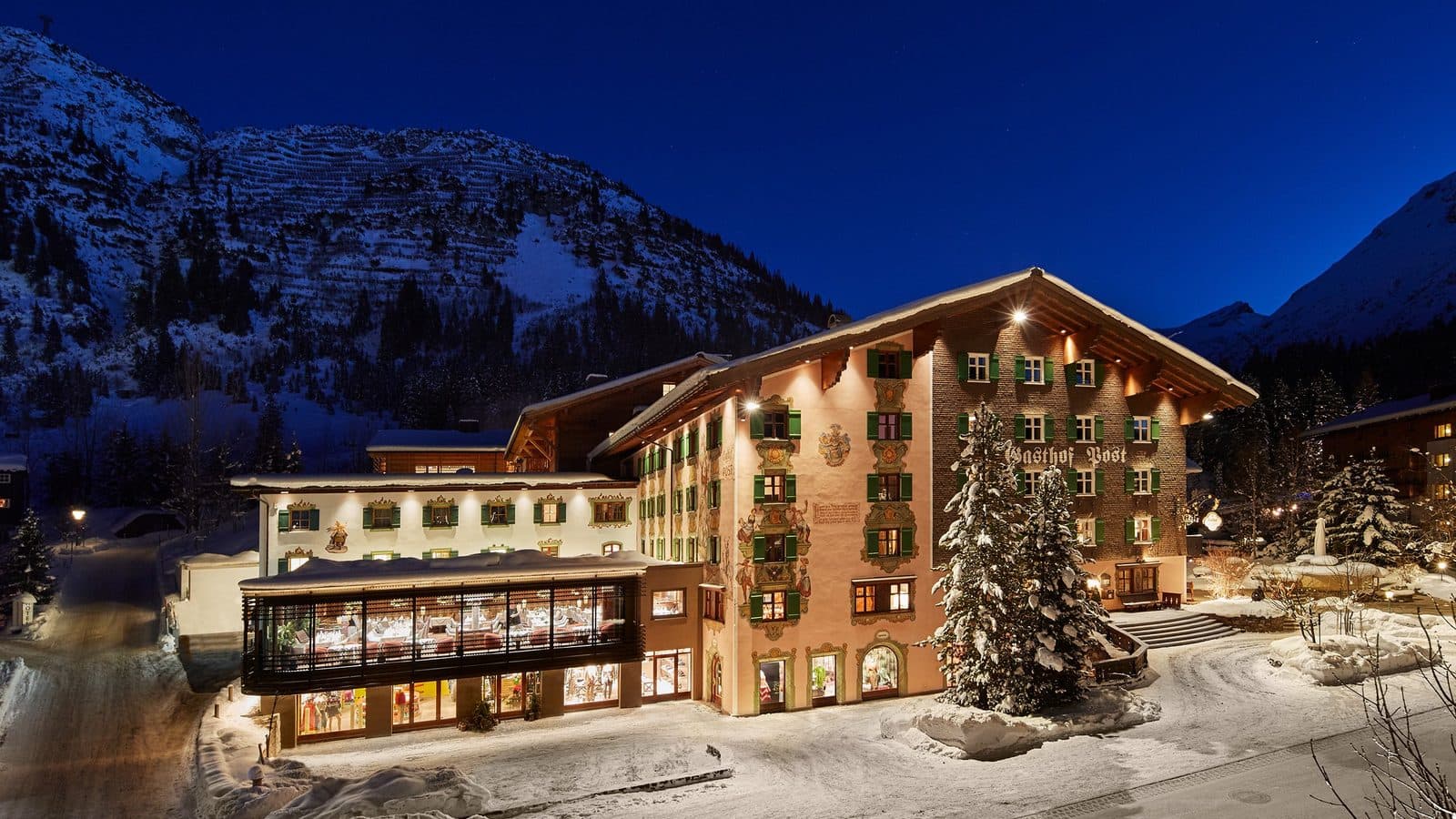 Hotel Post Lech вечером: освещённый фасад с зелёными ставнями, заснеженные ели и горы