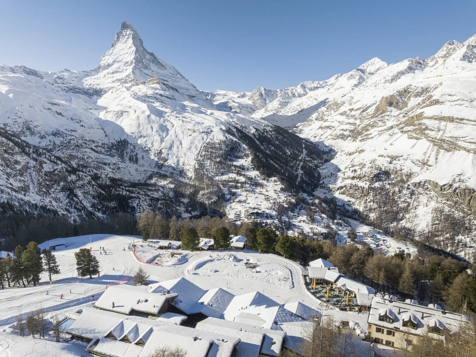 Riffelalp Resort зимой с высоты: заснеженные крыши отеля на плато среди лиственниц, за ним Маттерхорн и долина Церматта