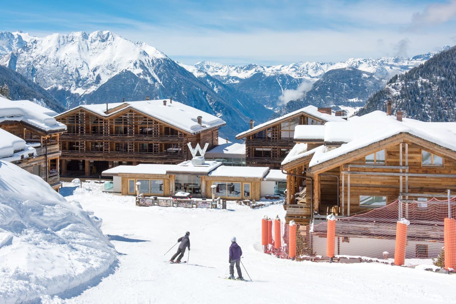 W Verbier in winter: wooden chalets with snowy roofs and a W sign, skiers on the piste in front and mountains behind