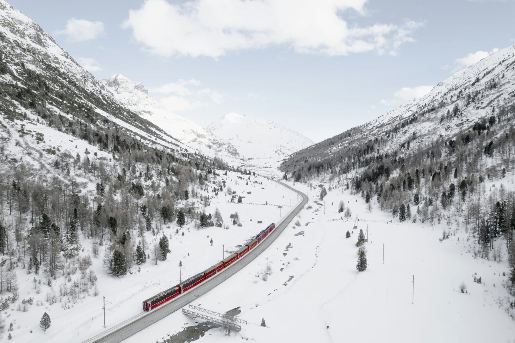A red Rhaetian Railway train crosses the snowy Bernina valley among larches, viewed from above