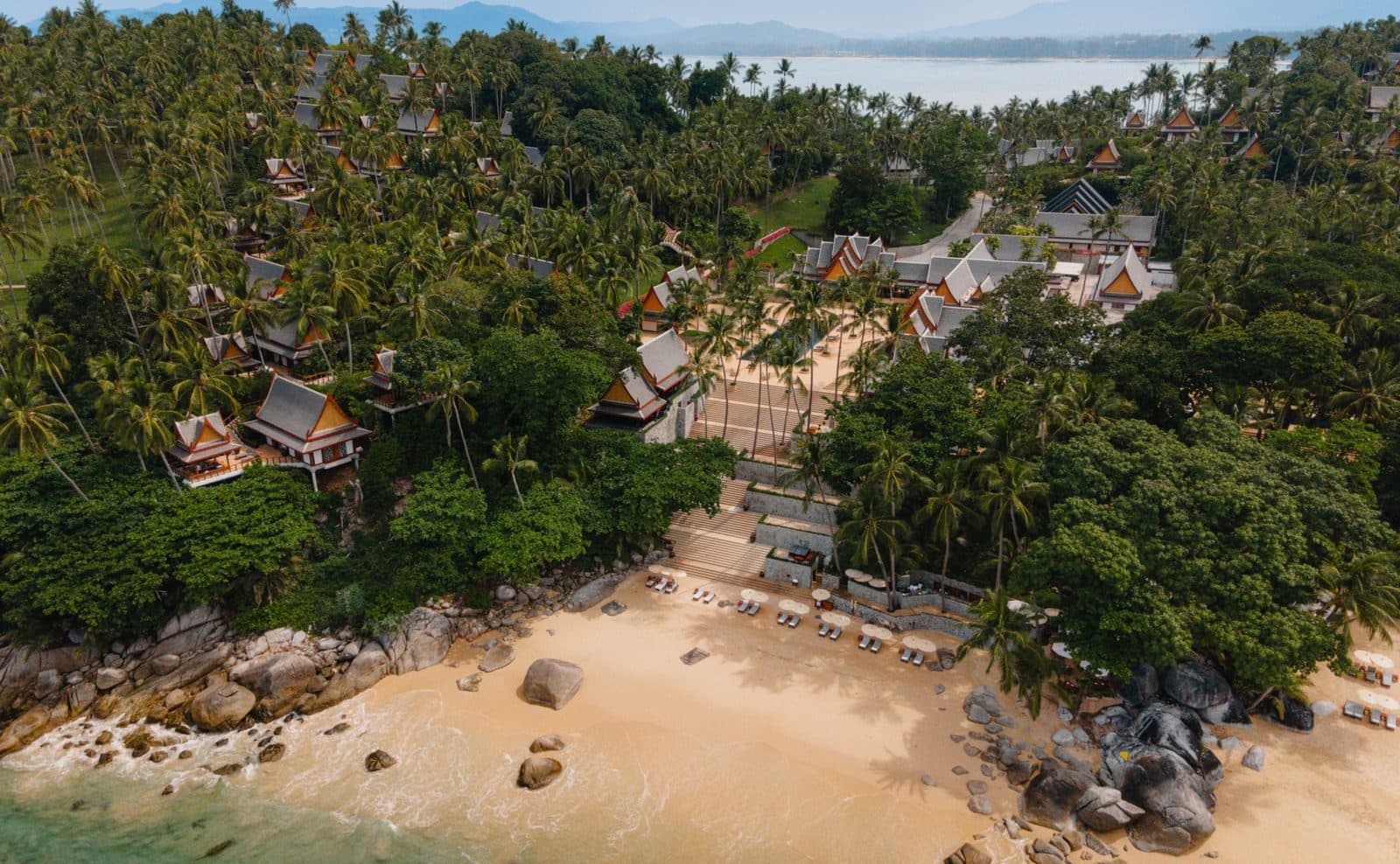 Aerial view of Amanpuri: Thai-roofed pavilions among coconut palms on the headland slope, Pansea Beach with loungers and waterside boulders