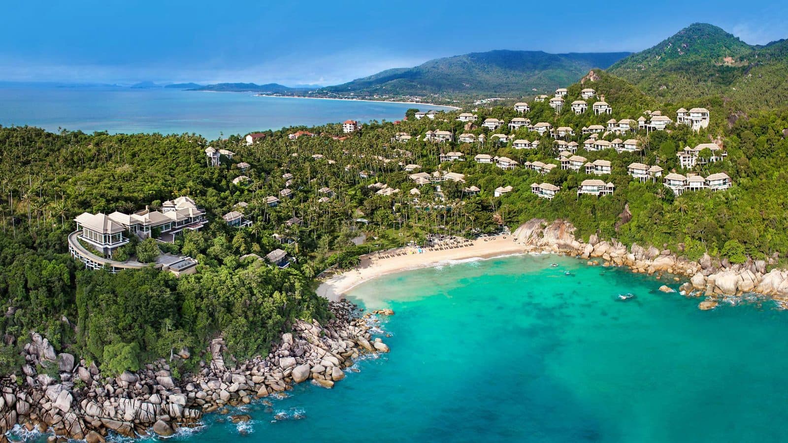 Aerial view of Banyan Tree Samui: white villas on a wooded slope above a beach bay, granite boulders by the water, with a long beach and mountains in the distance