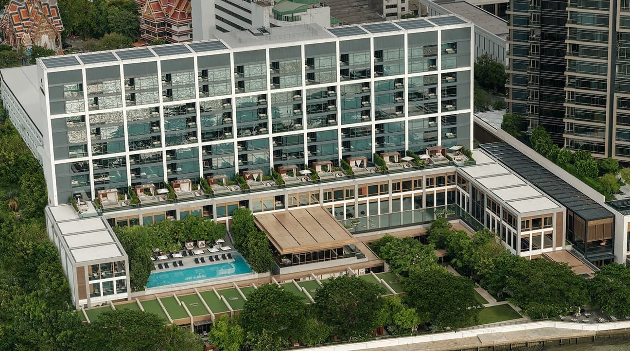 Вид сверху на Capella Bangkok: невысокое здание со стеклянным фасадом и террасами, бассейн и газоны в саду у набережной Чао Прайи, за ним видны крыши тайского храма