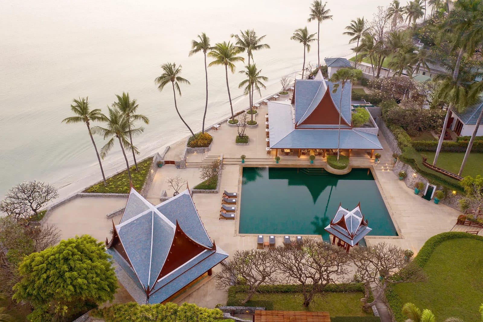 Aerial view of Chiva-Som: a seaside pool between Thai-roofed pavilions, loungers, coconut palms along the beach and calm Gulf of Thailand waters