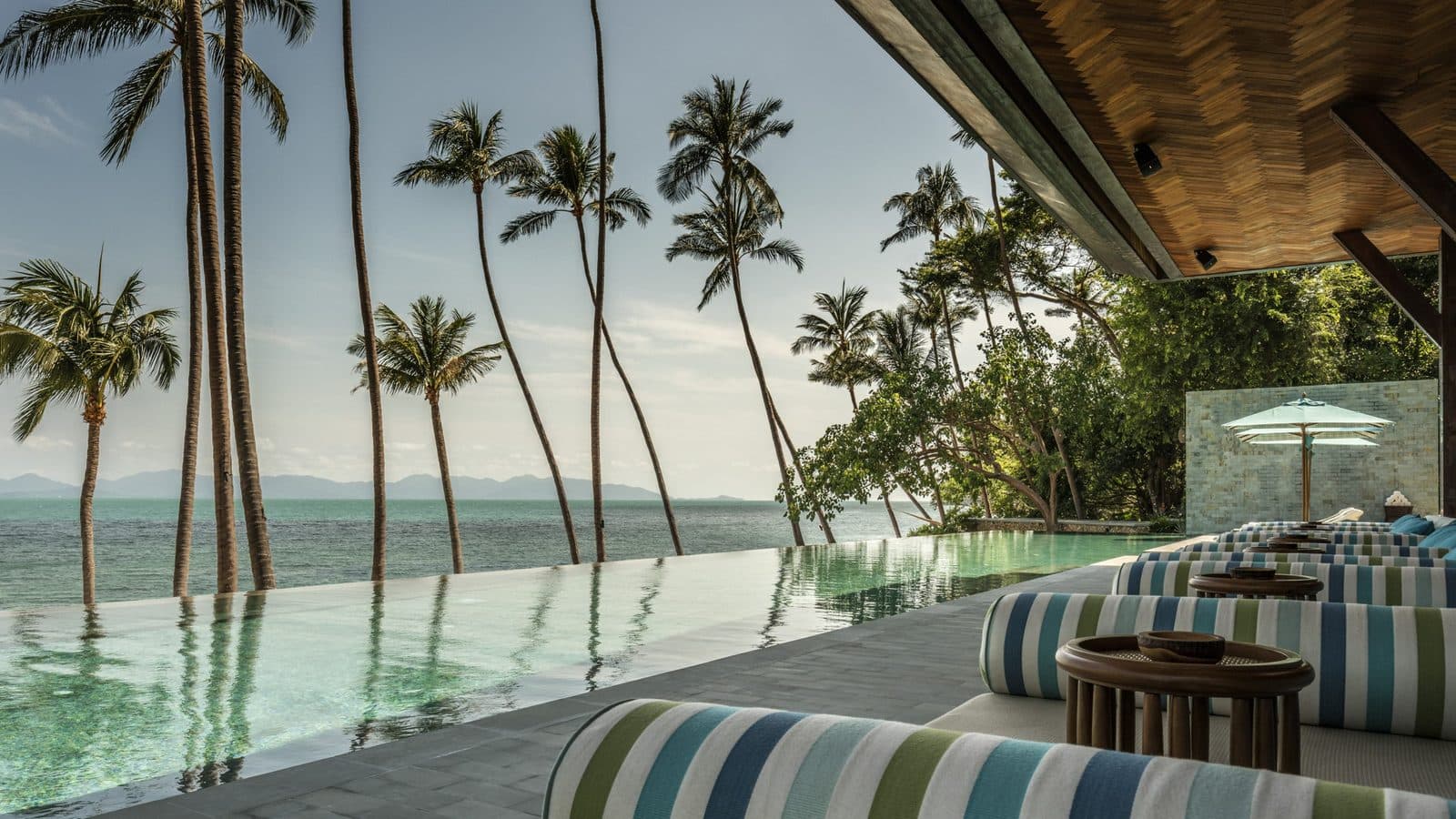 A seaside infinity pool framed by tall palms, striped loungers beneath a wooden canopy and the Gulf of Thailand to the horizon