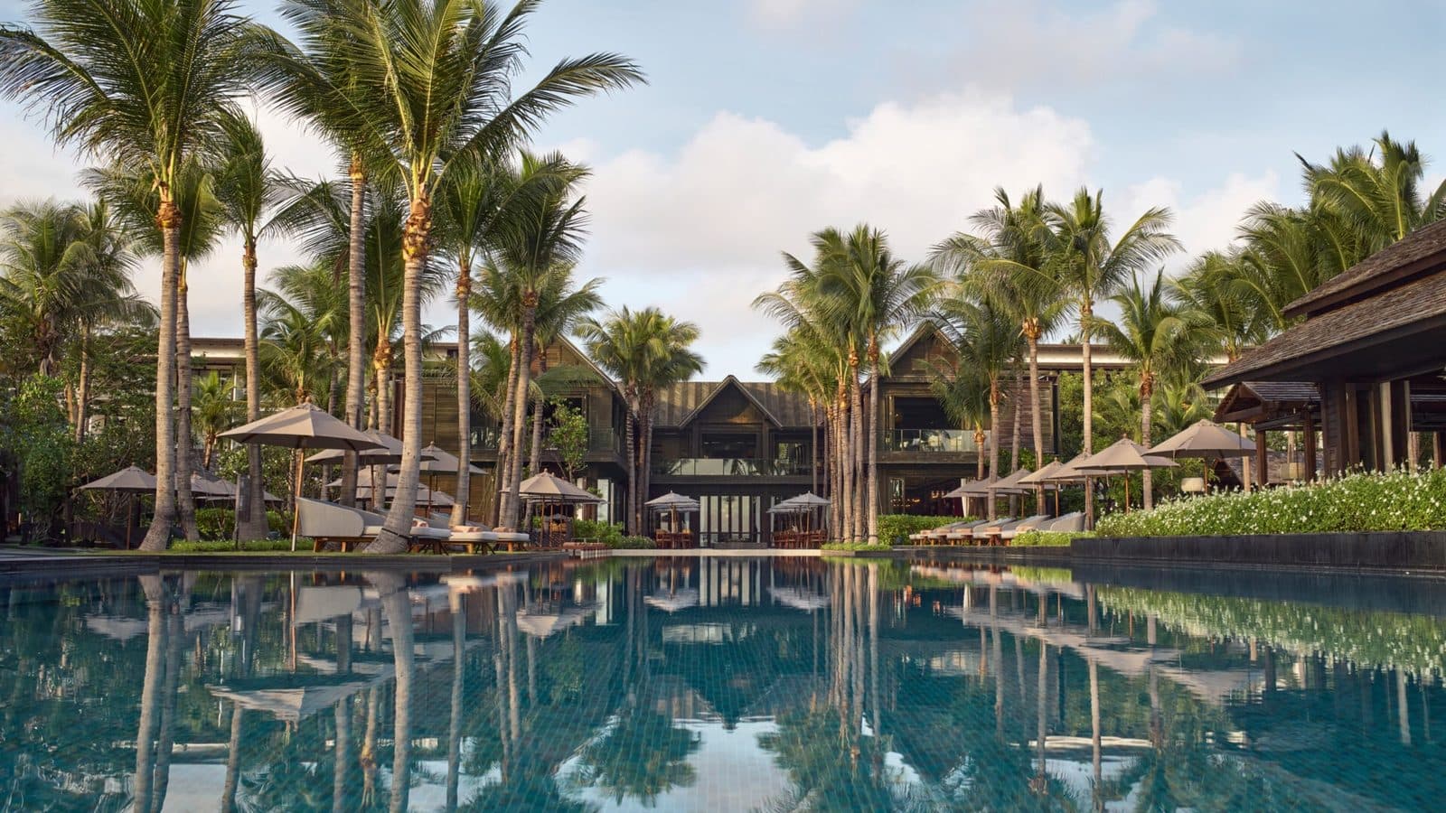 Главный бассейн Kimpton Kitalay Samui: в неподвижной воде отражаются кокосовые пальмы, зонтики и корпуса под тёмными скатными крышами
